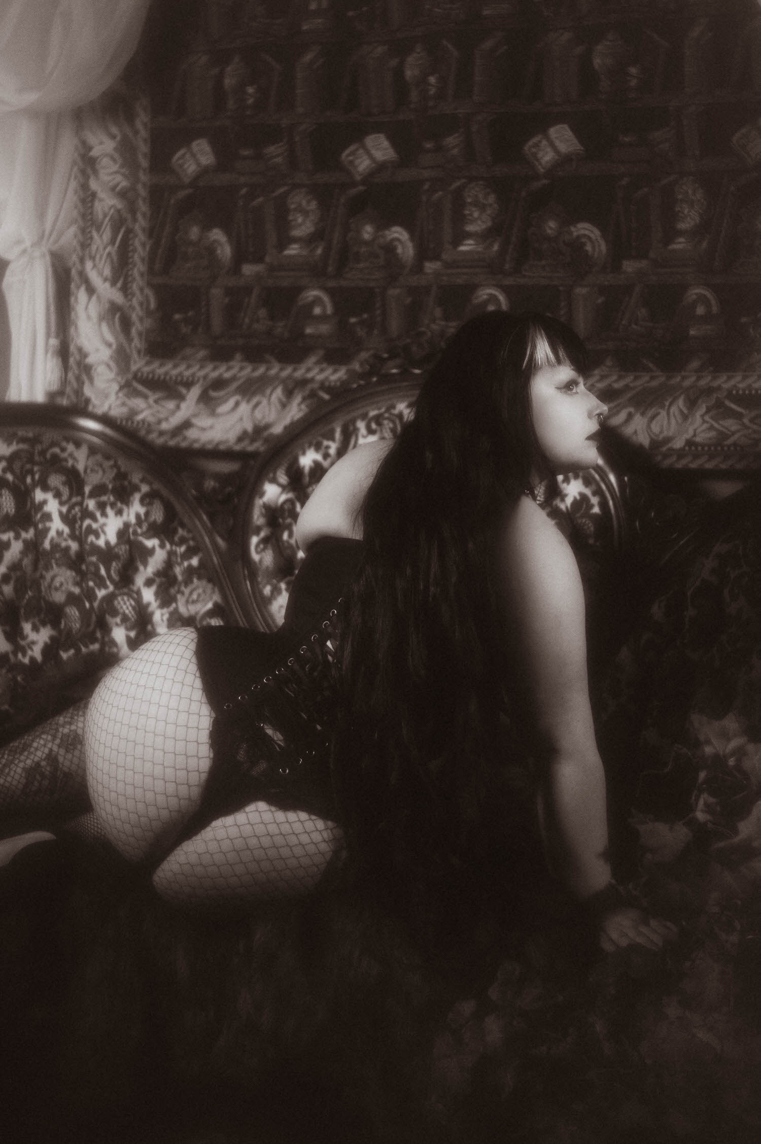 A black and white photo shows a bride kneeling on a patterned sofa in a black lace up corset and fishnet tights, looking off to the side during this black corset boudoir photoshoot, Royal Lune Photo.