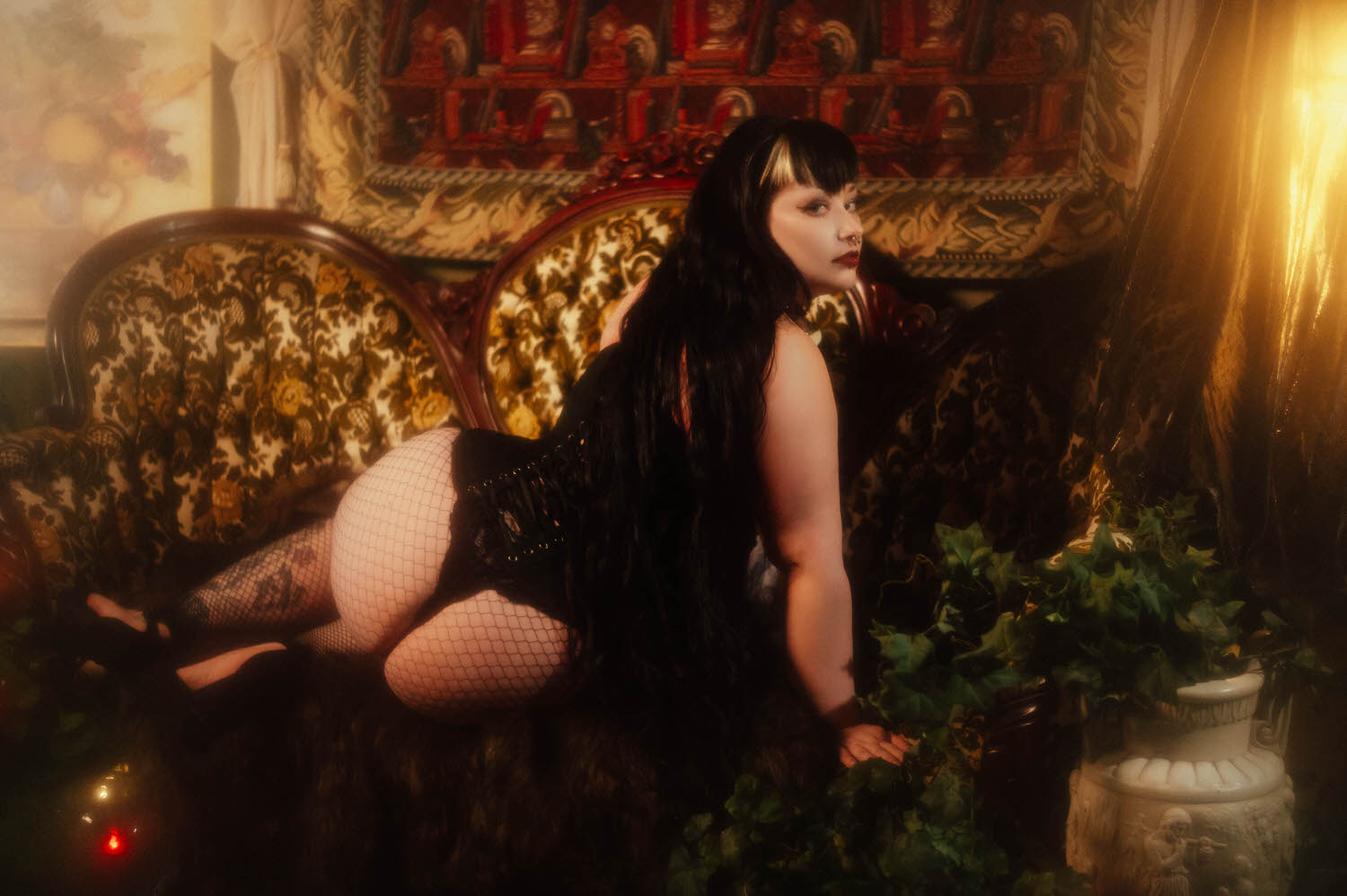 A bride kneels on a patterned Victorian sofa in a black corset and fishnet tights, looking back over her shoulder near a potted ivy plant for this gothic bridal boudoir session at Royal Lune Studio, Royal Lune Photo.