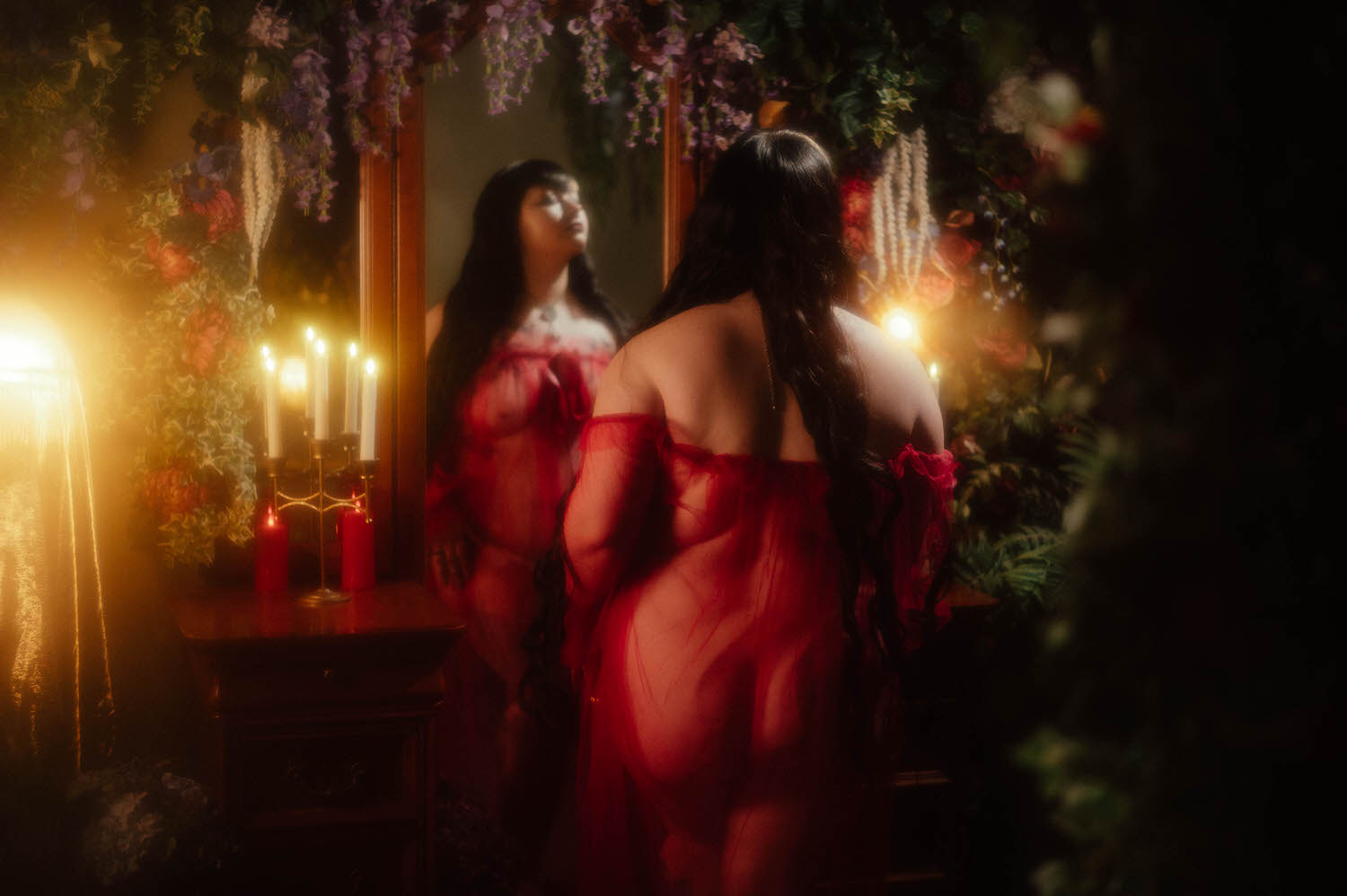 A bride in a sheer burgundy duster stands before an antique mirror reflecting her image, with white taper candles in a brass candelabra and red pillar candles on the vanity beside a wisteria and floral wall at Royal Lune Studio, Royal Lune Photo.