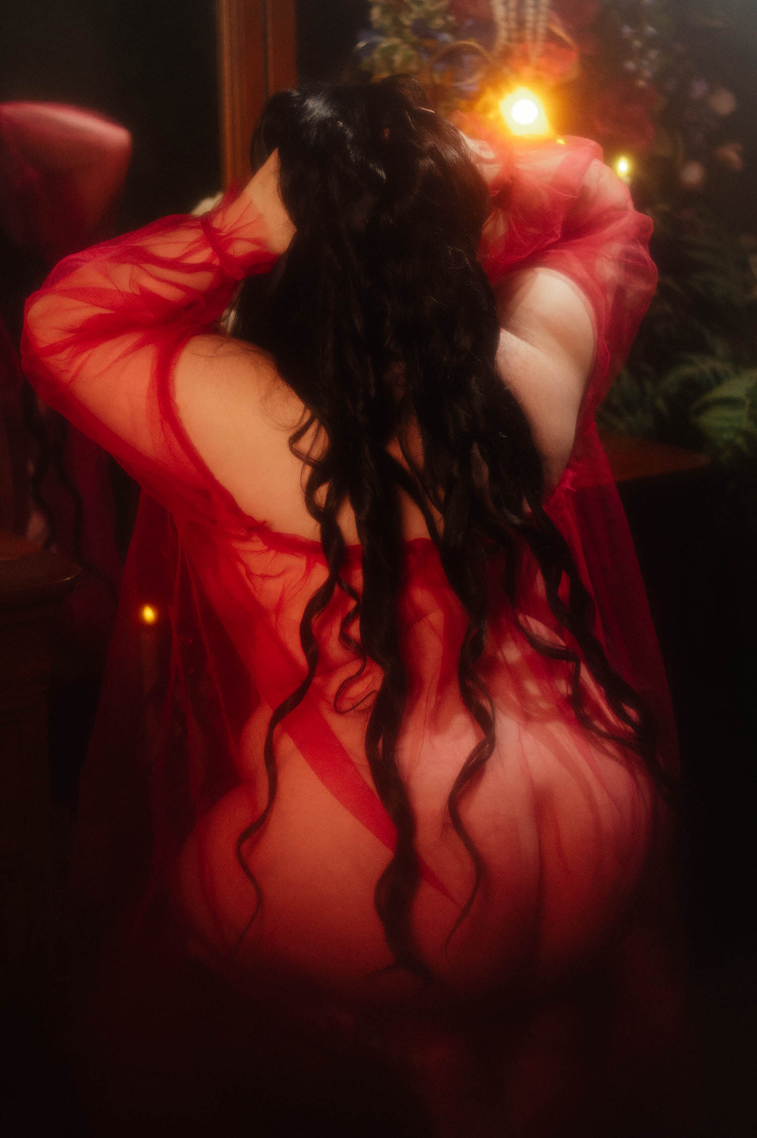 A bride gathers her long dark curled hair with both hands in a sheer burgundy duster, standing near a vanity with lit taper candles and a floral wall during this gothic bridal boudoir session at Royal Lune Studio, Royal Lune Photo.