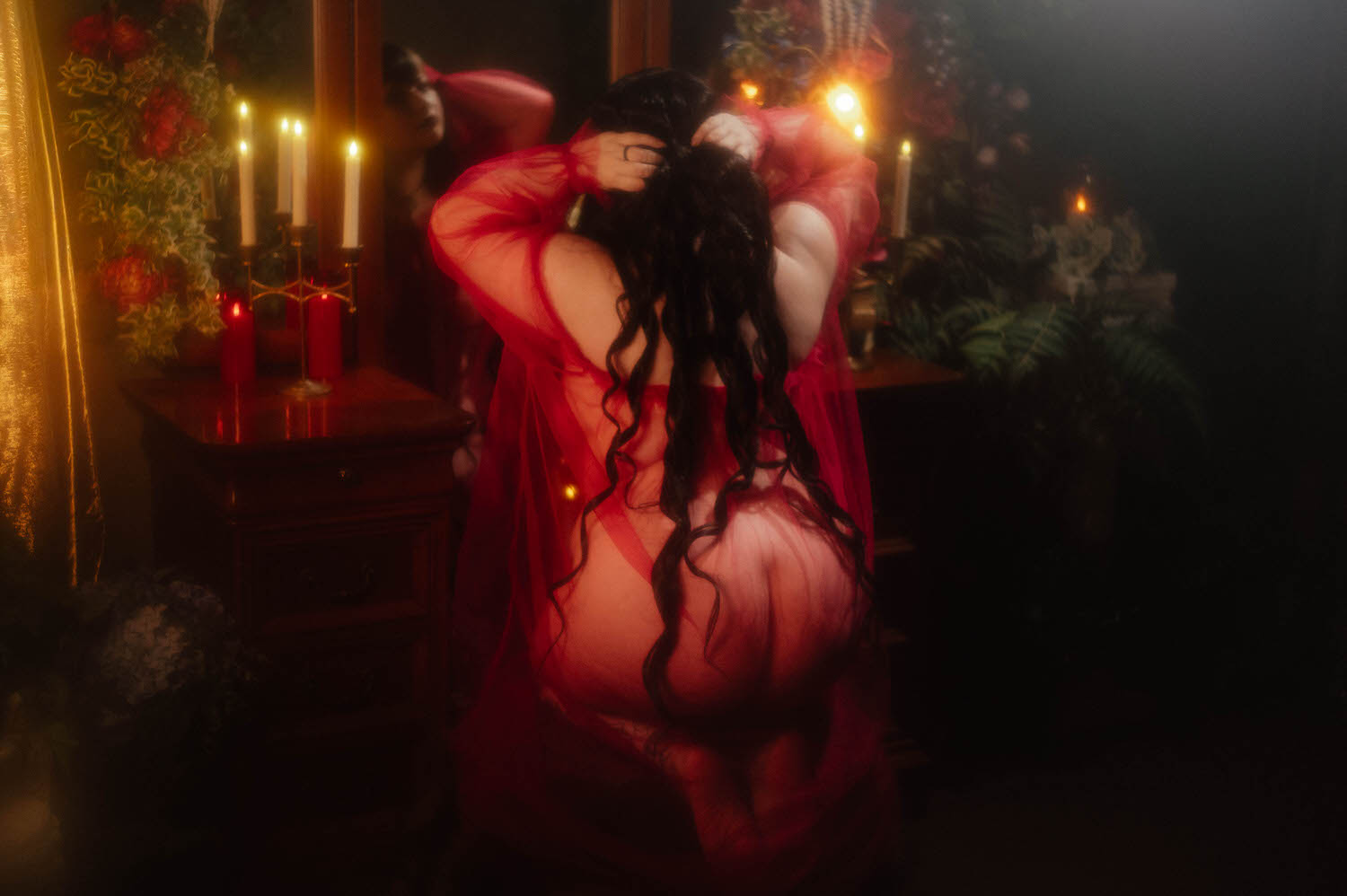 A bride lifts her long dark hair with both hands while wearing a sheer burgundy duster, photographed from behind against a wisteria and floral backdrop for this alternative bridal boudoir session, Royal Lune Photo.