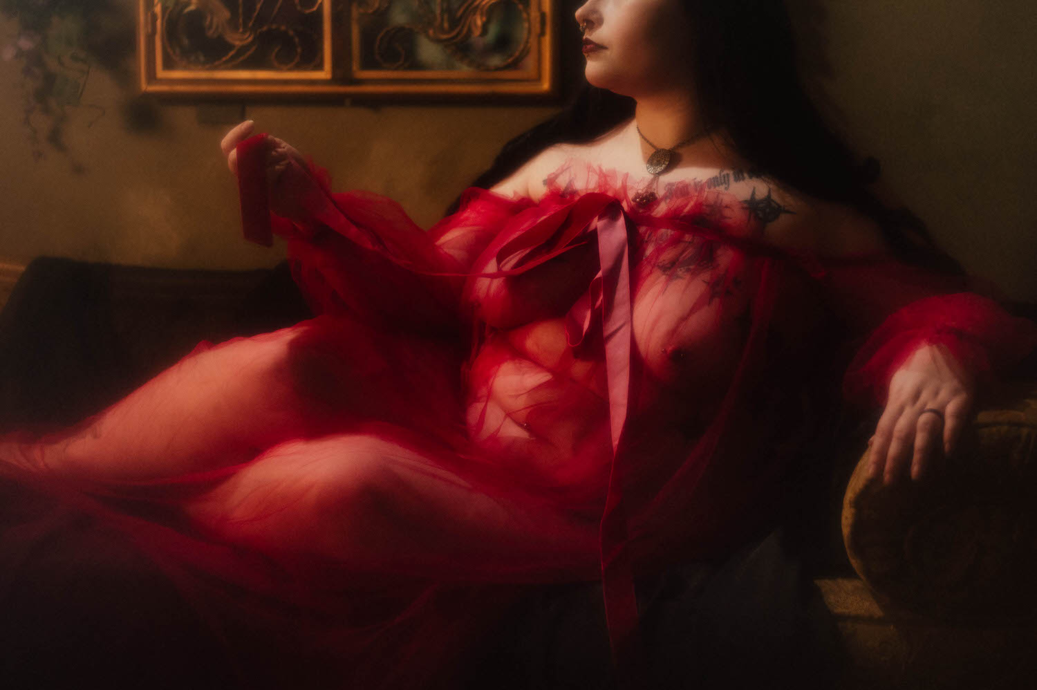 A close up shows a bride reclining on a tufted chaise in a sheer burgundy duster with a pendant necklace resting against her chest, framed by a wrought iron window piece during this gothic bridal boudoir session at Royal Lune Studio, Royal Lune Photo.