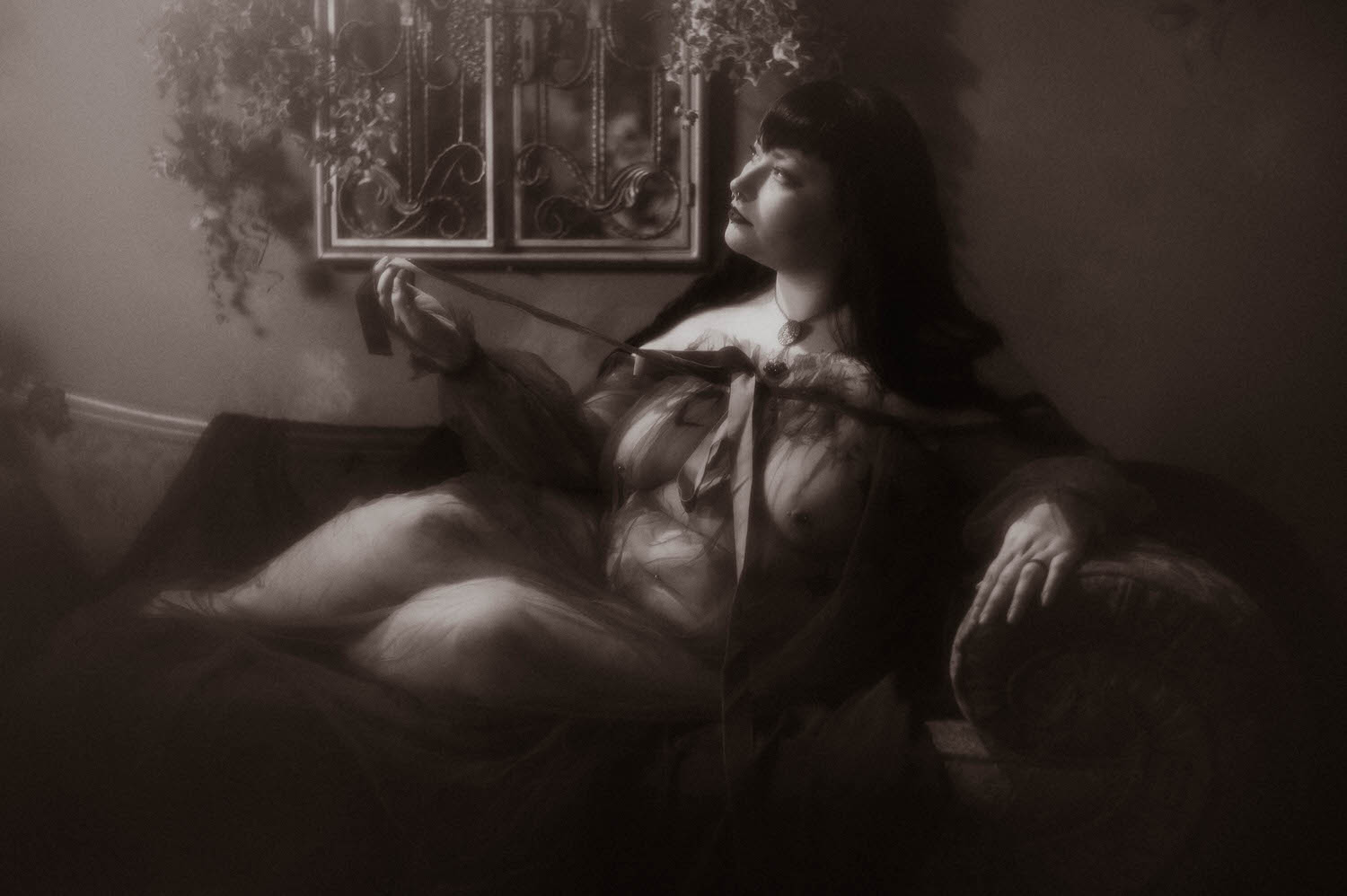 A black and white photo shows a bride reclining on a tufted chaise while pulling the velvet ribbon of her sheer duster, with a wrought iron window piece and trailing ivy on the wall behind her at Royal Lune Studio, Royal Lune Photo.