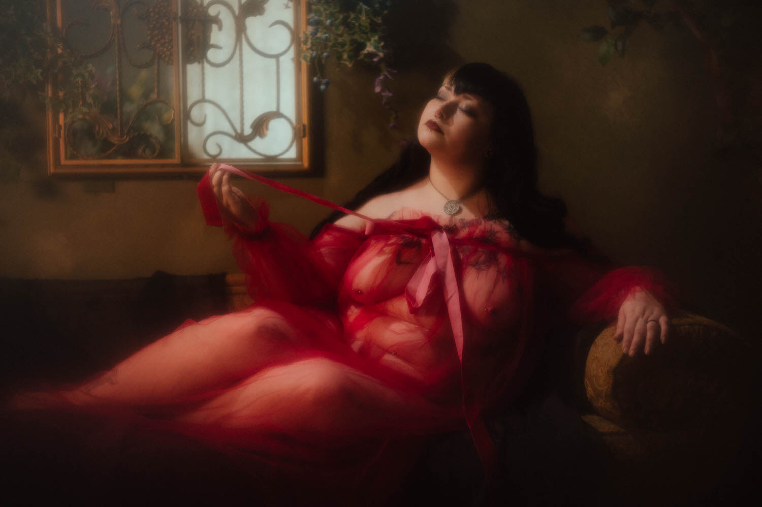 A bride reclines on a tufted chaise in a sheer burgundy duster, pulling the velvet ribbon at her chest beneath a wrought iron window piece and trailing ivy, for this alternative bridal boudoir session, Royal Lune Photo.