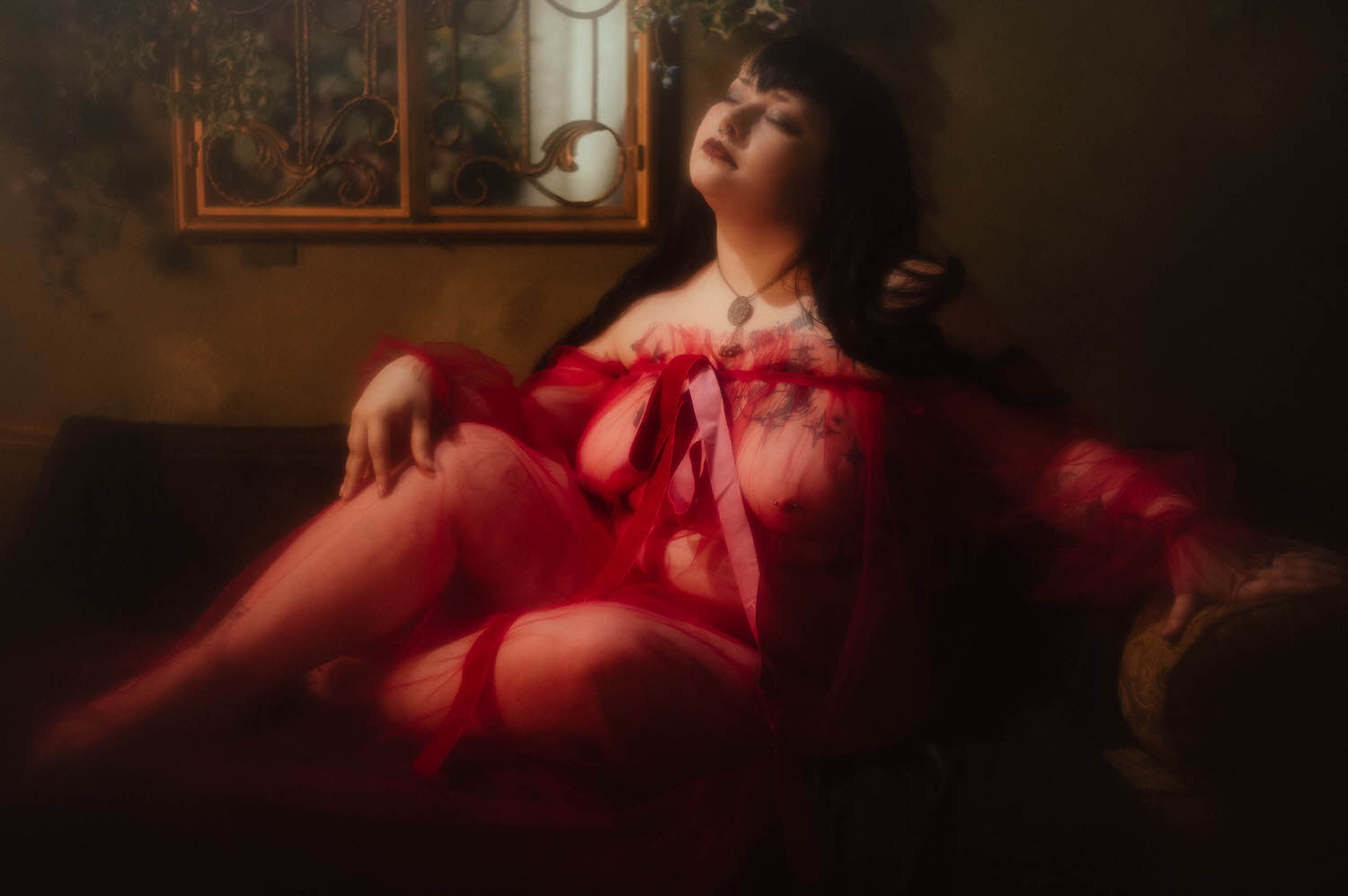 A bride reclines on a tufted chaise with her eyes closed and head tilted back, wearing a sheer burgundy duster beneath a wrought iron window piece at Royal Lune Studio, Royal Lune Photo.