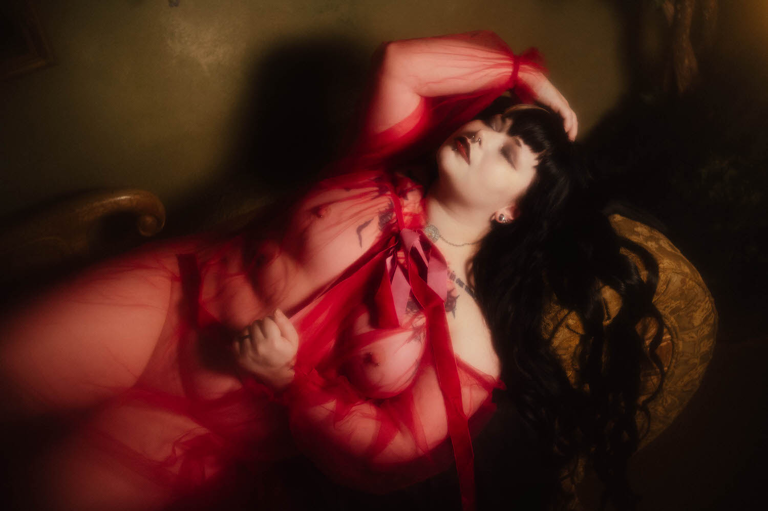 An overhead shot shows a bride pulling the velvet ribbon of her sheer burgundy duster with her dark hair spread across a gold patterned cushion for this gothic bridal boudoir session, Royal Lune Photo.