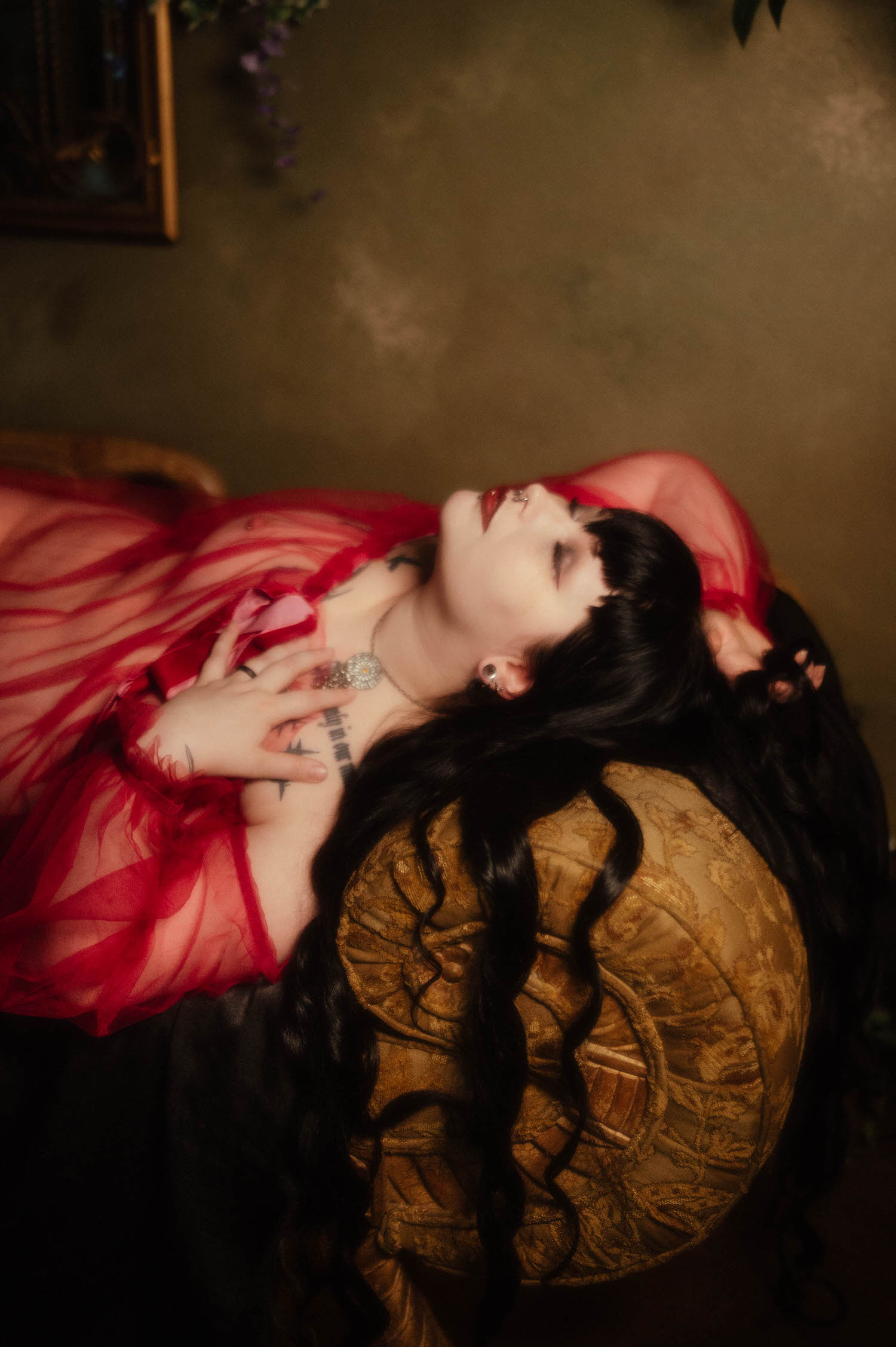 A wide overhead shot shows a bride stretched across a gold patterned chaise in a sheer burgundy duster with her dark hair fanned out beneath her for this alternative bridal boudoir session, Royal Lune Photo.