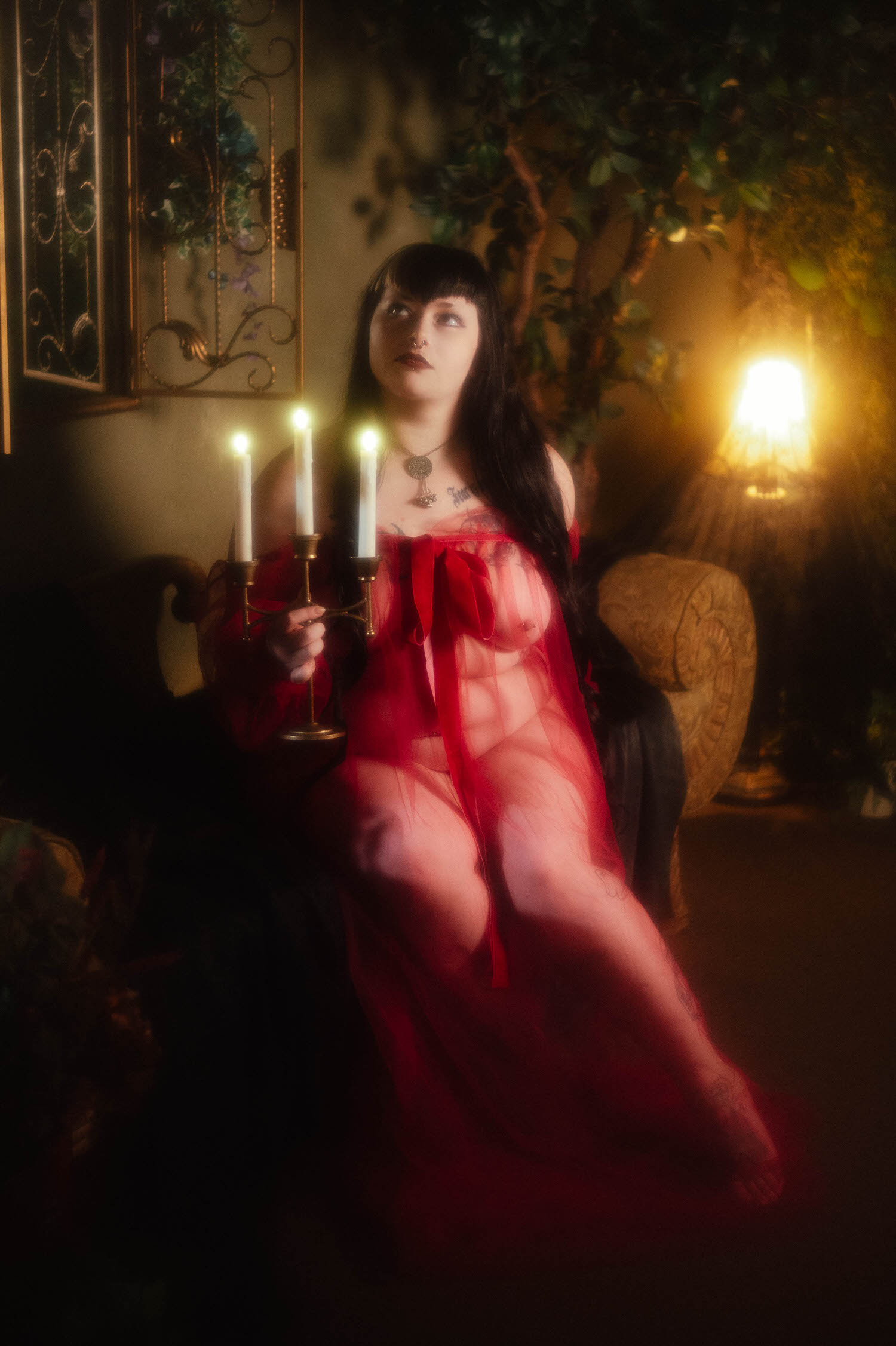 A bride sits upright holding a brass candelabra with three lit taper candles, wearing a sheer burgundy duster beside a potted tree and glowing lamp at Royal Lune Studio, photographed by bridal boudoir photographer Dallas Fort Worth, Royal Lune Photo.