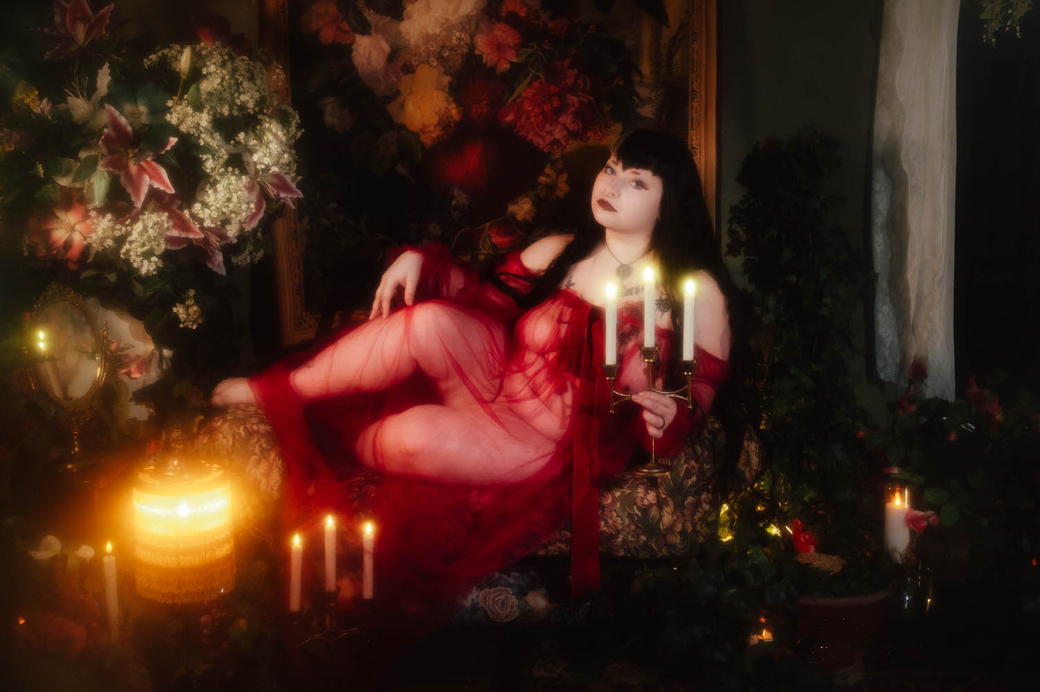 A bride sits among white lilies, baby's breath, and lit candles while holding a brass candelabra in a sheer burgundy duster during this alternative bridal boudoir session at Royal Lune Studio, Royal Lune Photo.