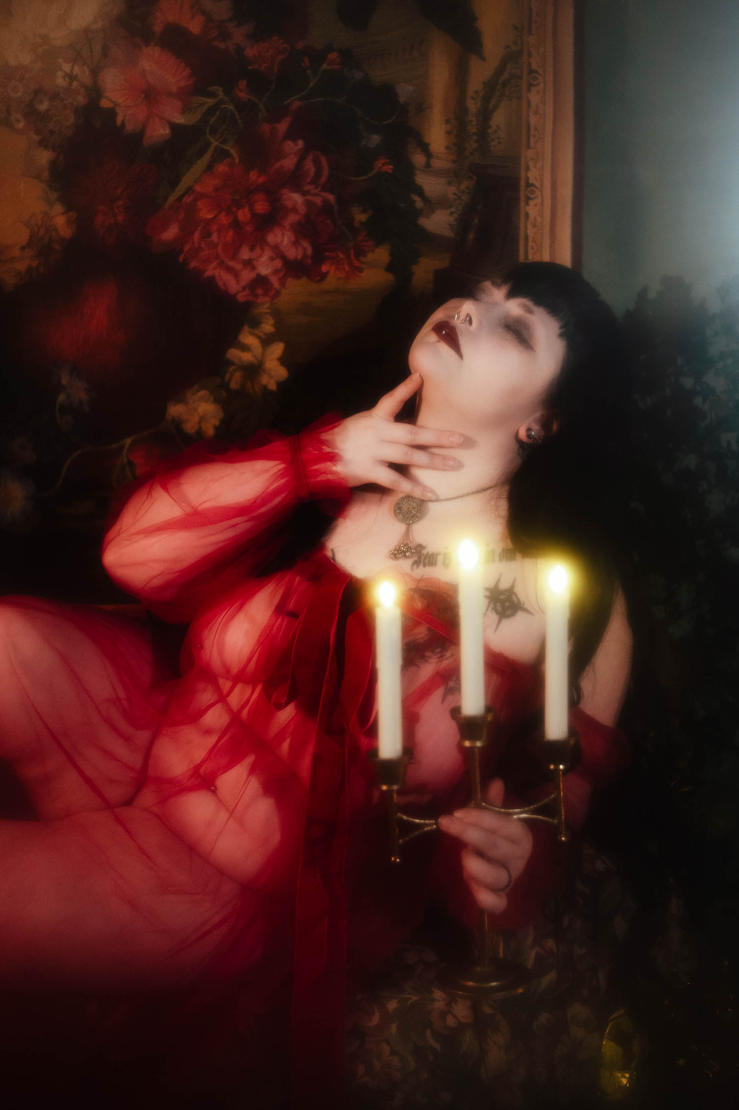 A bride reclines against a floral tapestry backdrop with her hand resting at her throat, wearing a sheer burgundy duster beside three lit white taper candles in a brass candelabra, photographed by bridal boudoir photographer Dallas Fort Worth, Royal Lune Photo.