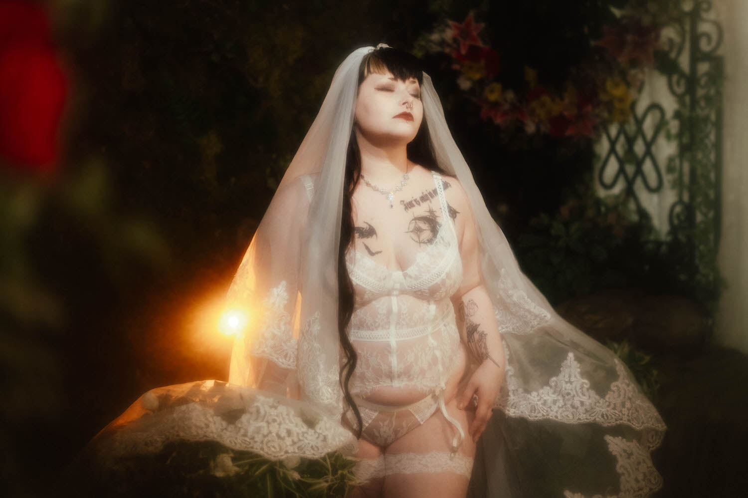 A bride stands in a white lace bridal lingerie set with a cathedral veil swept back off her face, lit by a single warm light beside a wrought iron trellis for this bridal boudoir with a wedding veil, Royal Lune Photo.