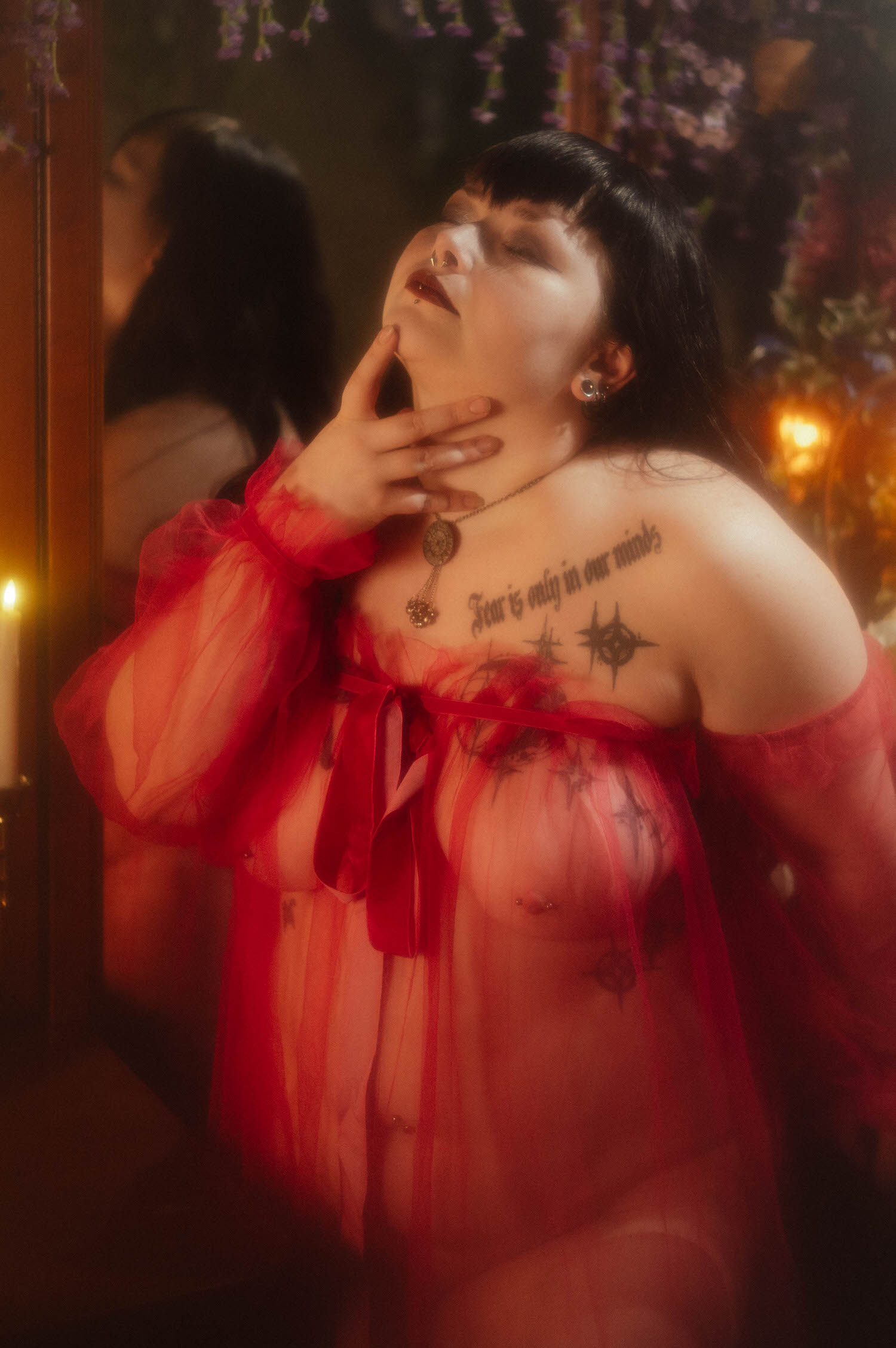 A close up portrait shows a bride with her head tilted back and her hand resting on her neck, wearing a sheer burgundy duster beside a single lit candle and a floral backdrop, for this alternative bridal boudoir session, Royal Lune Photo.