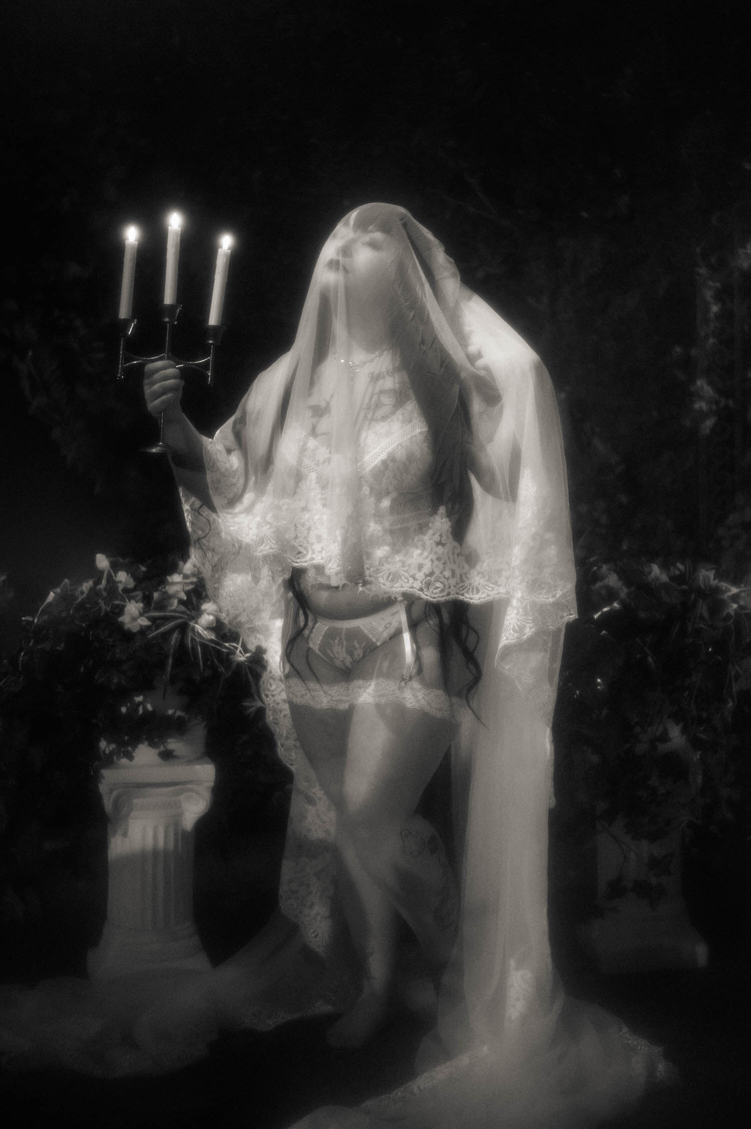 A black and white photo shows a bride holding a lit candelabra with her veil pulled forward over her face, standing beside a stone pedestal dressed in white florals in a white lace bridal set, Royal Lune Photo.