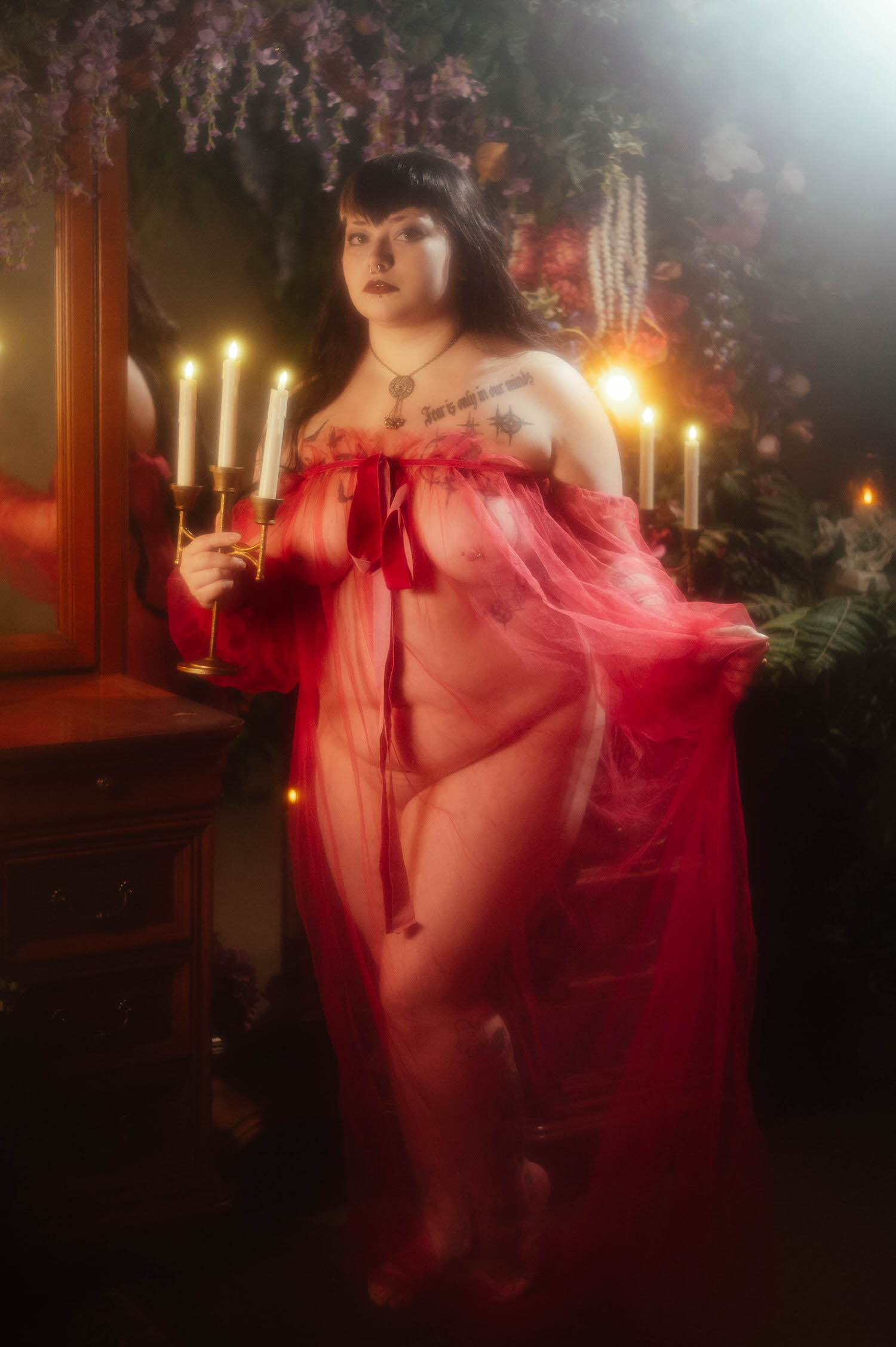 A bride poses in a sheer burgundy duster while holding a lit taper candelabra beside an antique vanity and mirror, framed by a wisteria and floral wall at Royal Lune Studio, Royal Lune Photo.