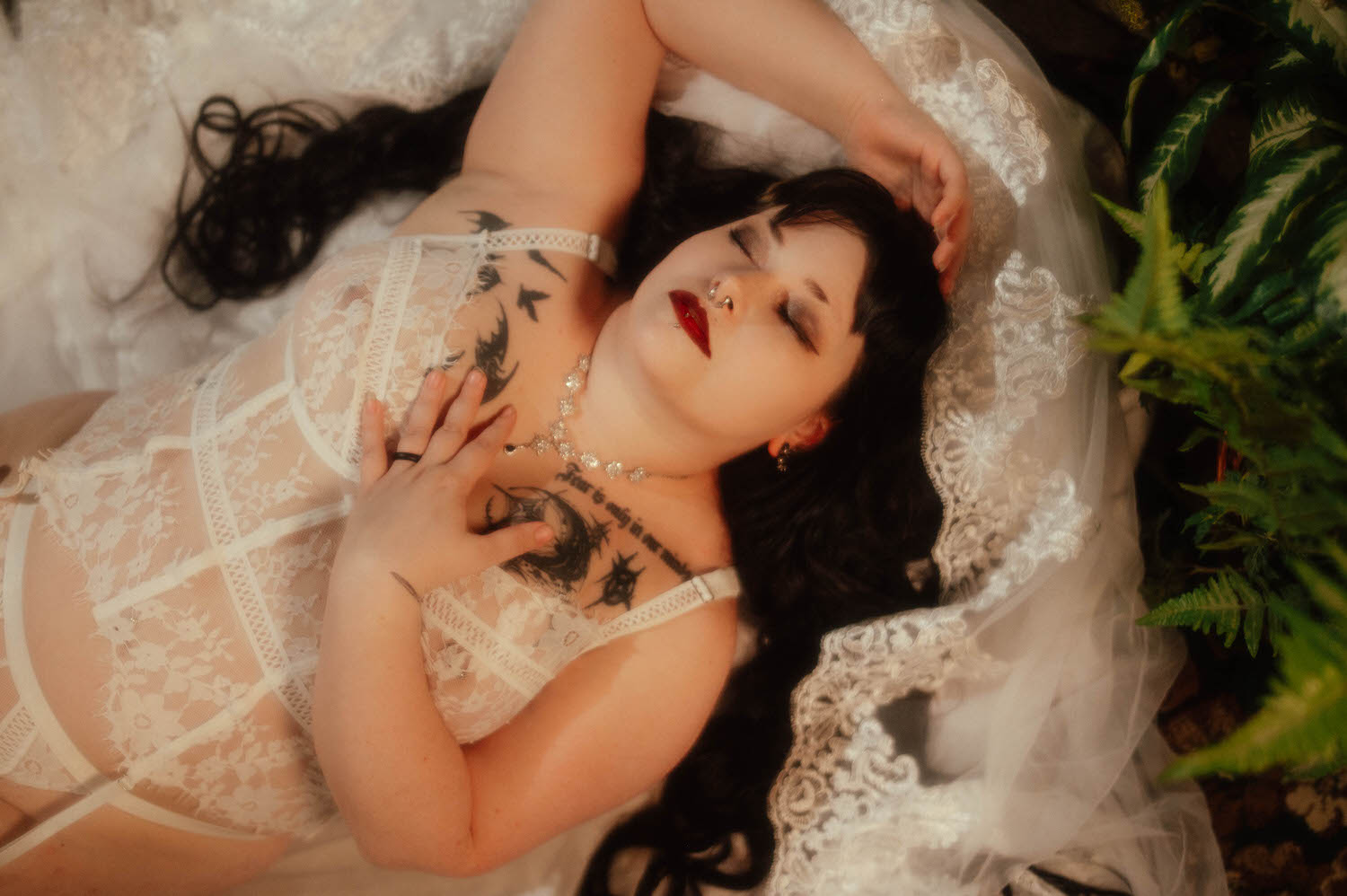 An overhead shot shows a bride lying on lace veil fabric with her hand resting on her chest tattoos, wearing a white lace bridal bra and garter set beside a fern for this bridal boudoir gift for your fiancée, Royal Lune Photo.