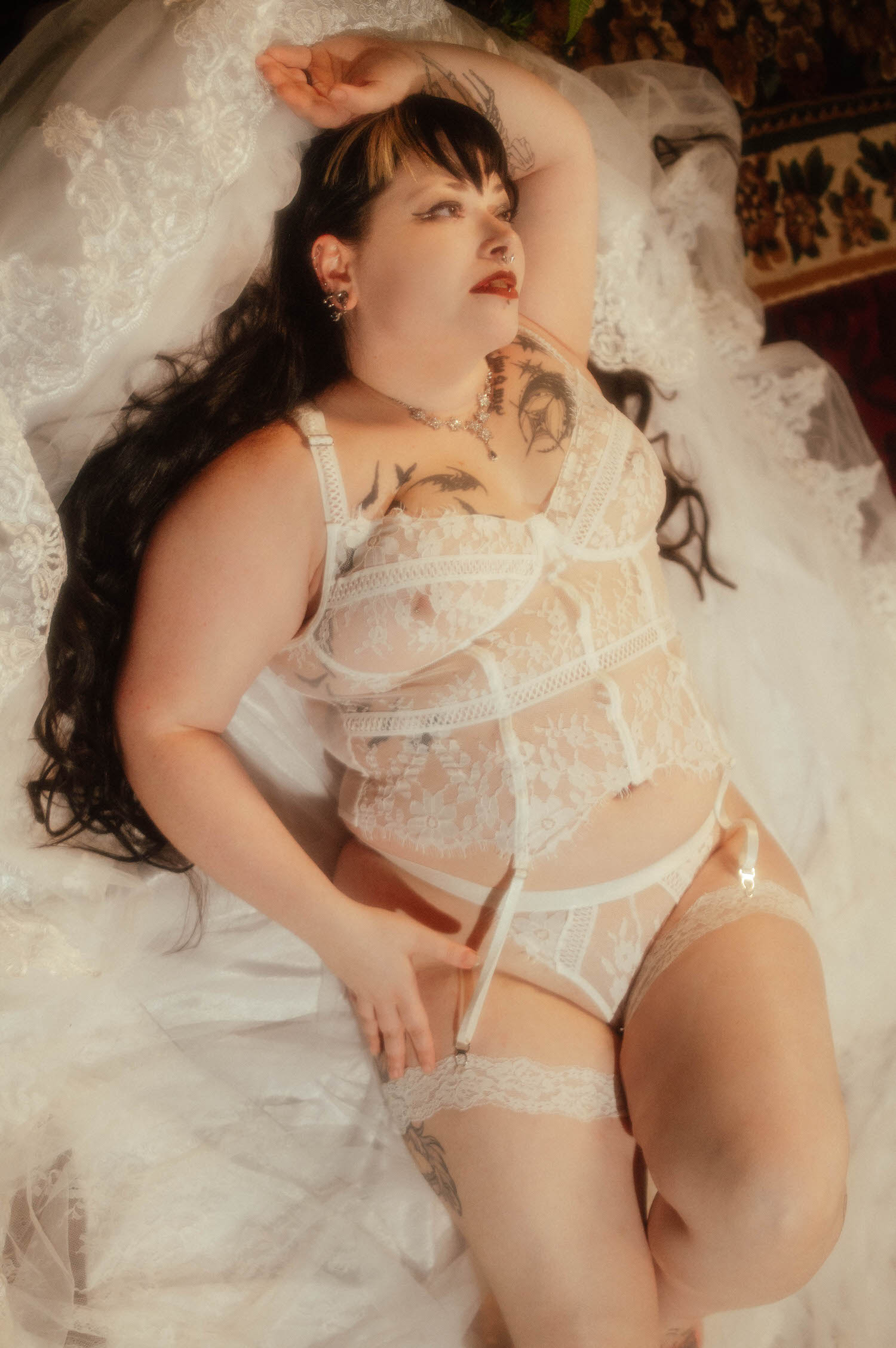 A bride lies among lace veil fabric with one arm raised overhead, wearing a white lace bra and garter set with a garter clip visible at her thigh during this gothic bridal boudoir session at Royal Lune Studio, Royal Lune Photo.