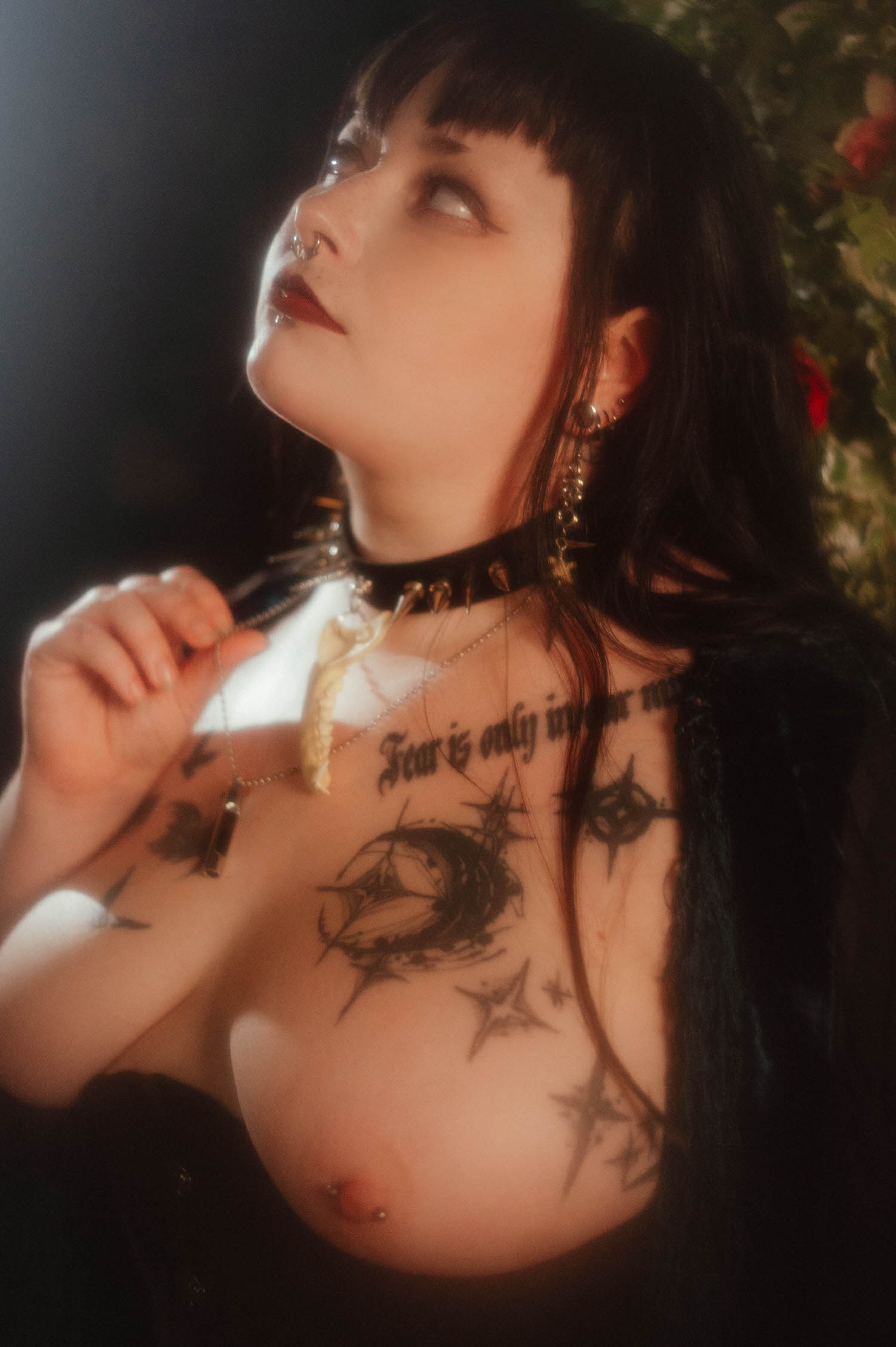A close up shows a bride holding a bone shaped pendant on a spiked choker necklace against her tattooed chest for this black corset boudoir photoshoot, Royal Lune Photo.