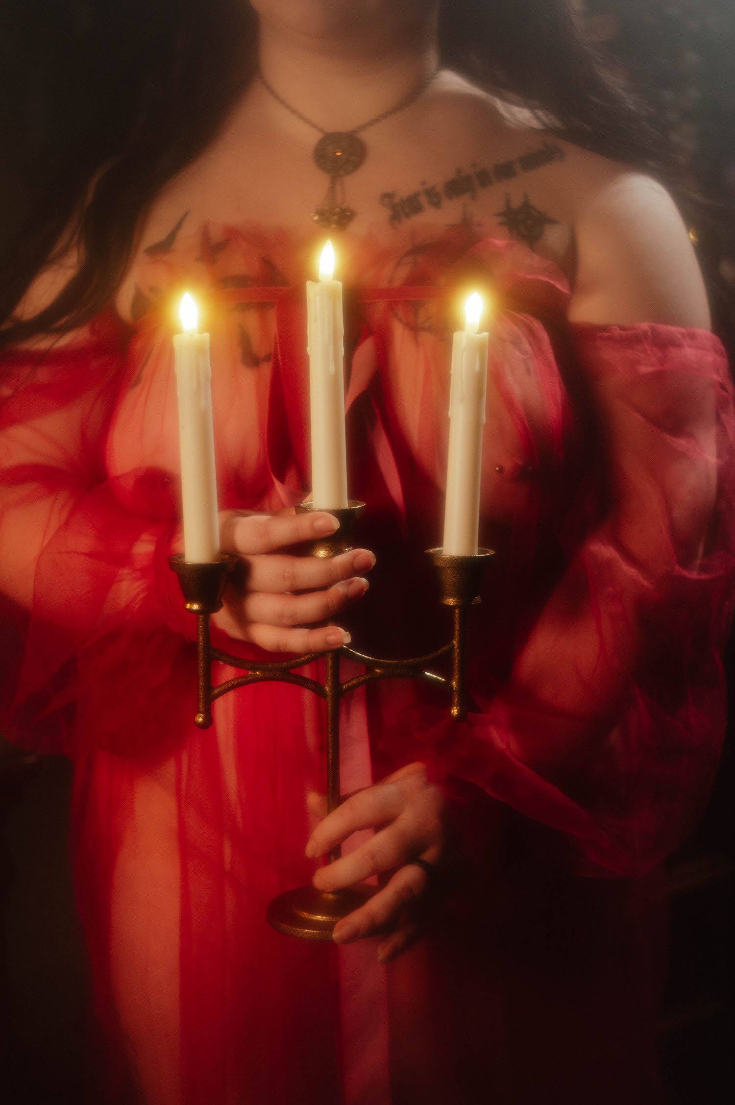 A close up shows a bride in a sheer burgundy duster holding a brass candelabra with three lit taper candles against her chest during a gothic bridal boudoir session, Royal Lune Photo.