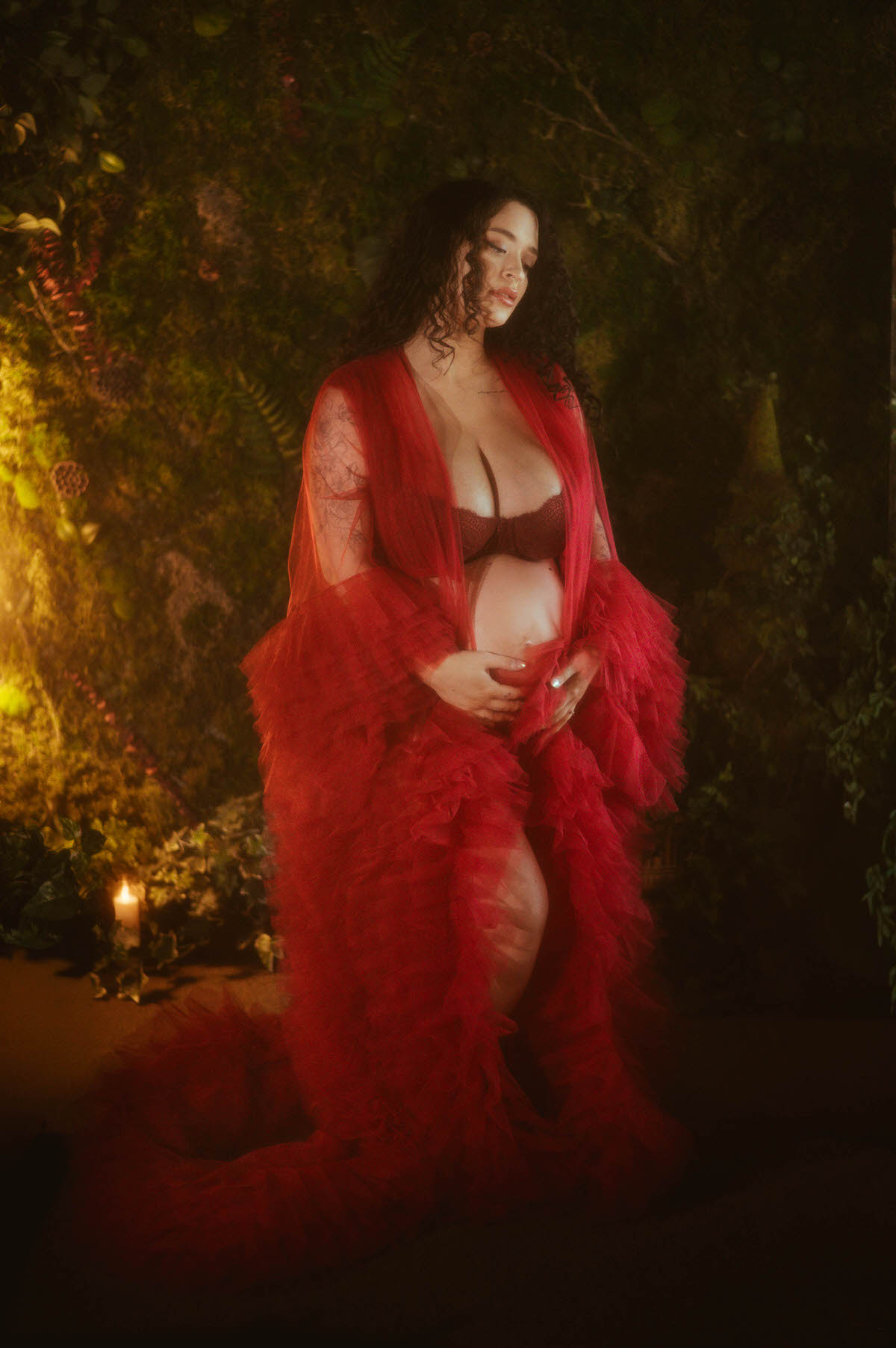 An artistic maternity photoshoot Dallas session captures a pregnant woman standing in a voluminous red sheer tulle robe worn open over a matching dark red lace bra, lit by warm candlelight against a lush mossy backdrop at Royal Lune Photo.