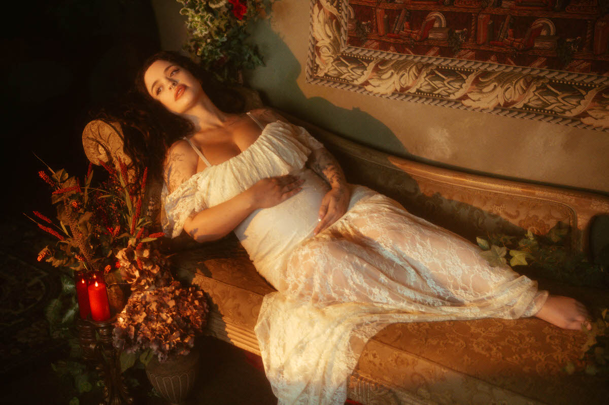 This maternity portrait studio Dallas image shows a woman reclining on a tapestry backed chaise in an ivory lace gown, looking toward the camera with one hand resting on her belly beside red pillar candles at Royal Lune Photo.