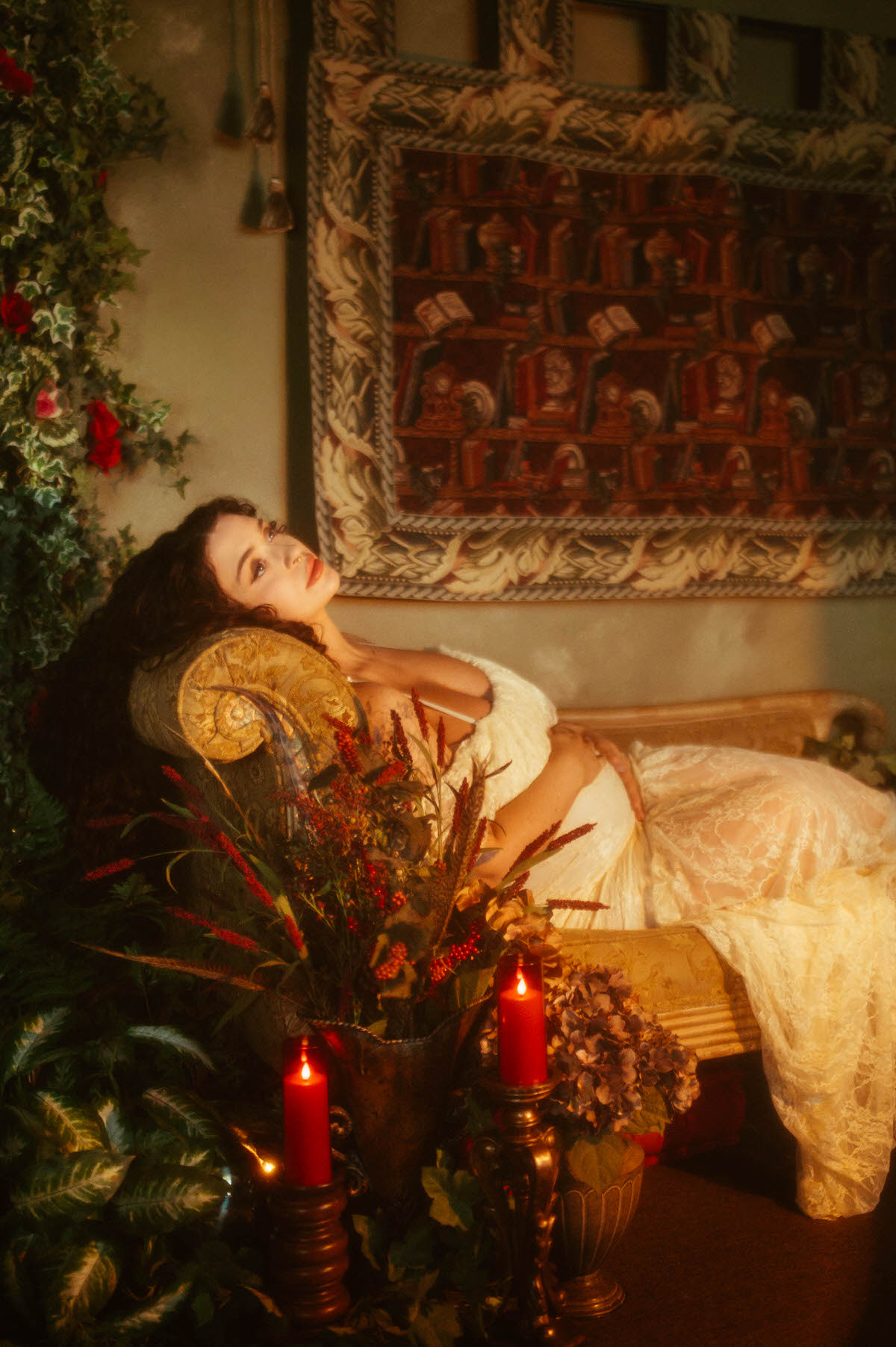 A Dallas maternity photographer captures a woman reclining on a tapestry backed chaise in an ivory lace gown, head tilted back with eyes closed, flanked by red pillar candles and a floral arrangement at Royal Lune Photo.