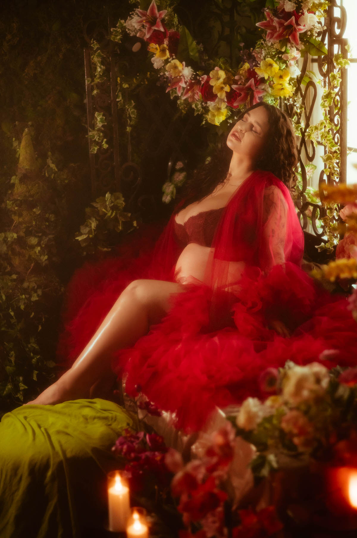 An artistic maternity photoshoot Dallas session features a pregnant woman seated among pink and yellow florals in a wrought iron alcove, eyes closed and legs stretched across a green vintage fabric cushion, wrapped in a flowing red tulle robe at Royal Lune Photo.