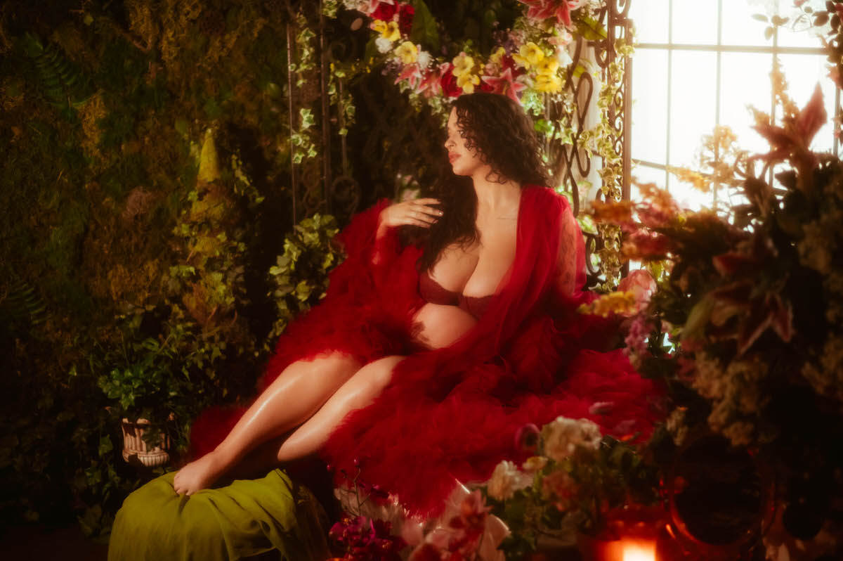 A moody maternity photography portrait shows a pregnant woman with her eyes open looking off to the side, one hand raised near her collarbone, reclining in a red tulle robe surrounded by pink and yellow blooms at Royal Lune Photo.