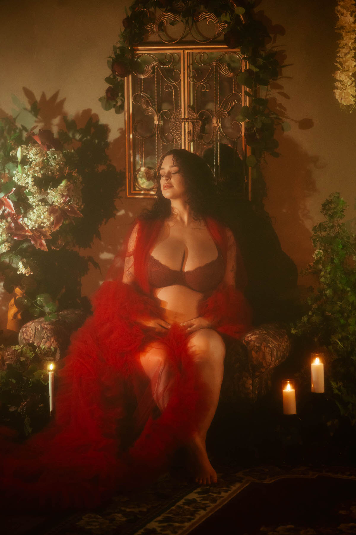 An artistic maternity photoshoot Dallas image shows a woman seated in a floral upholstered armchair, eyes closed, wearing a red sheer tulle robe over dark red lace lingerie in front of an antique gold framed mirror flanked by pillar candles at Royal Lune Photo.