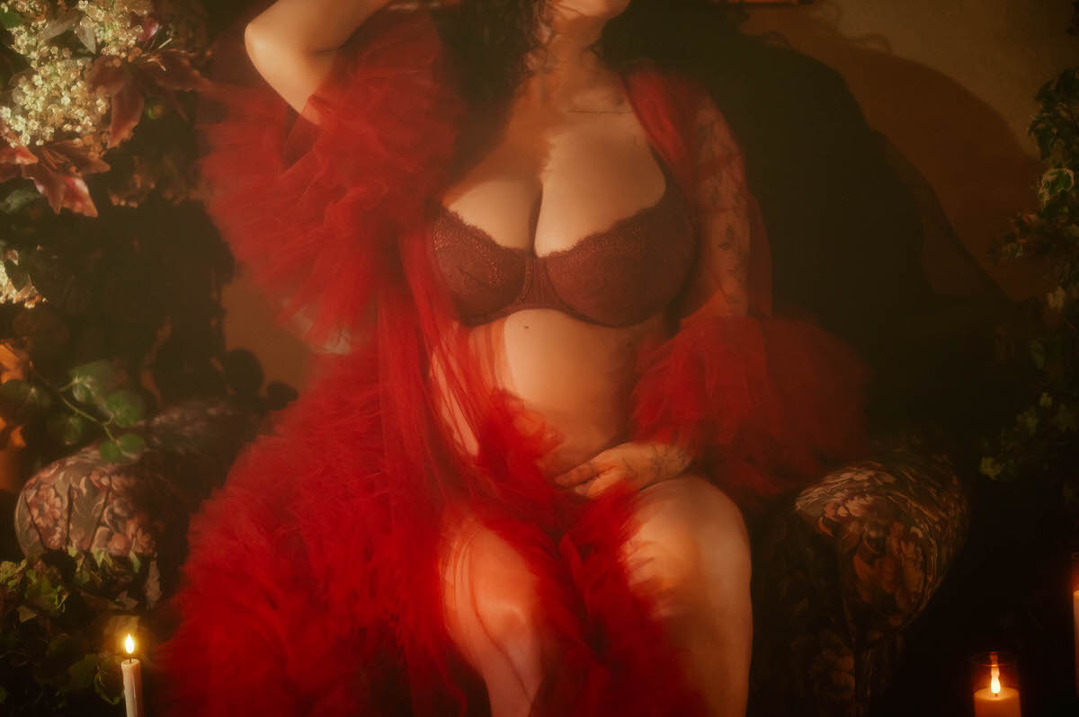 A close up maternity portrait studio Dallas image shows a pregnant belly and dark red lace bra beneath a sheer red tulle robe, with one hand resting on the bump while seated in a floral upholstered armchair at Royal Lune Photo.
