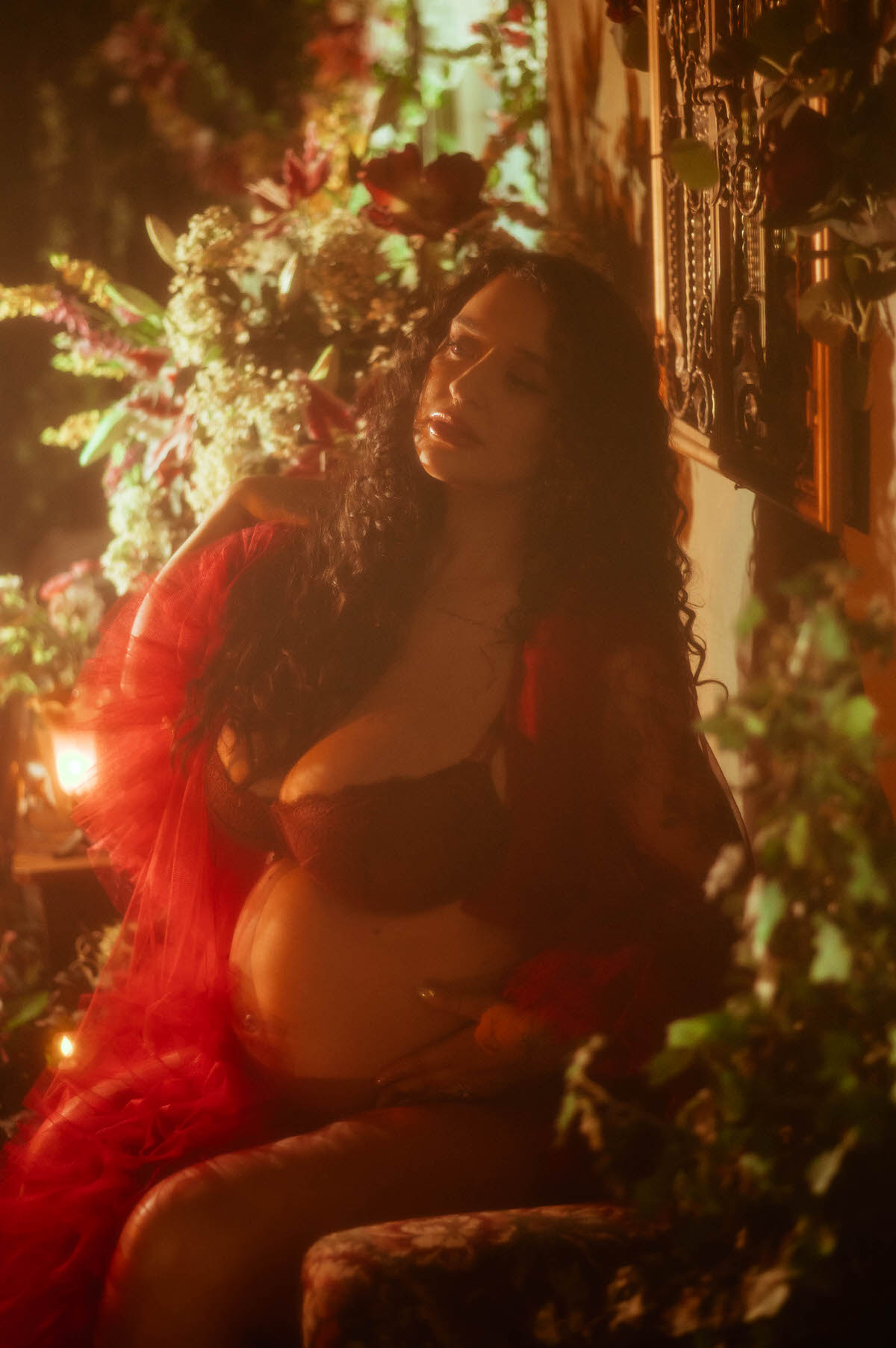 A moody maternity photography portrait captures a woman with her eyes closed and one arm raised behind her head, wrapped in red sheer tulle and surrounded by ivory and blush florals at Royal Lune Photo.