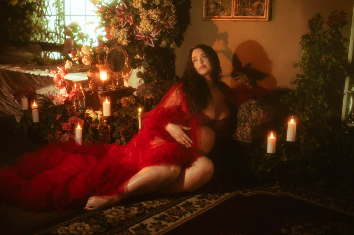 These pregnancy photoshoot ideas include a woman seated on a patterned rug with red tulle pooling around her, looking off to the side, lit by a row of pillar candles and soft window light at Royal Lune Photo.