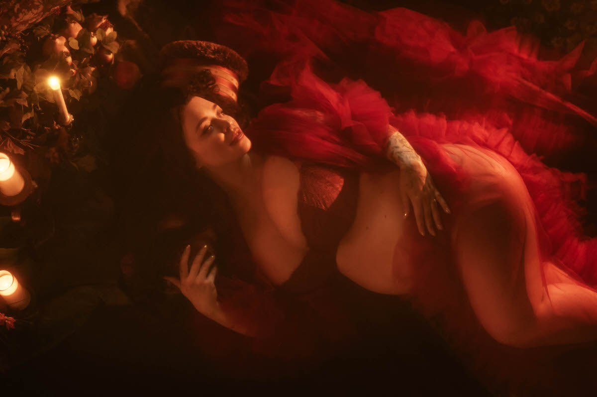 An artistic maternity photoshoot Dallas image captures an overhead view of a woman reclining in red sheer tulle against a patterned pillow for support, with one hand resting near her collarbone and floral shoulder tattoo work visible at Royal Lune Photo.