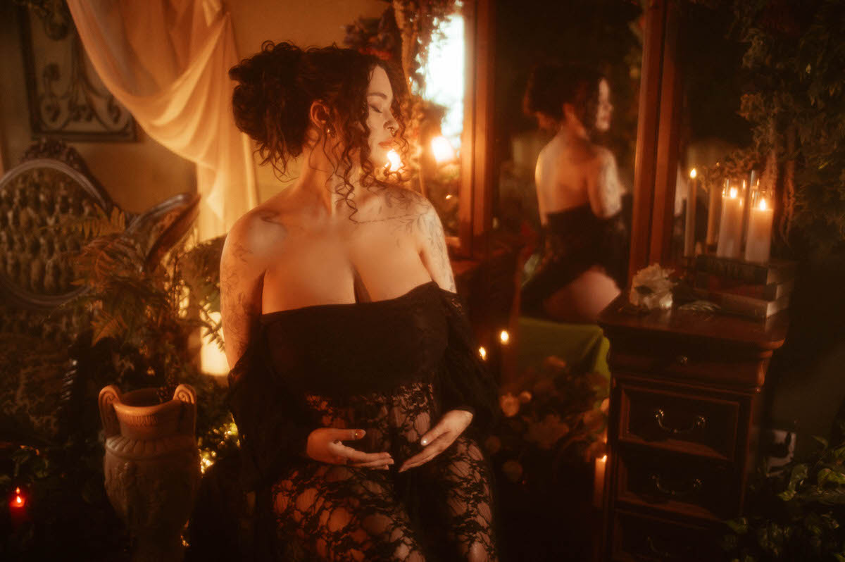 An artistic maternity photoshoot Dallas session features a woman with her hair styled in an updo, hands clasped in front of her black lace gown, standing near an antique urn with her reflection visible in a mirror behind her at Royal Lune Photo.