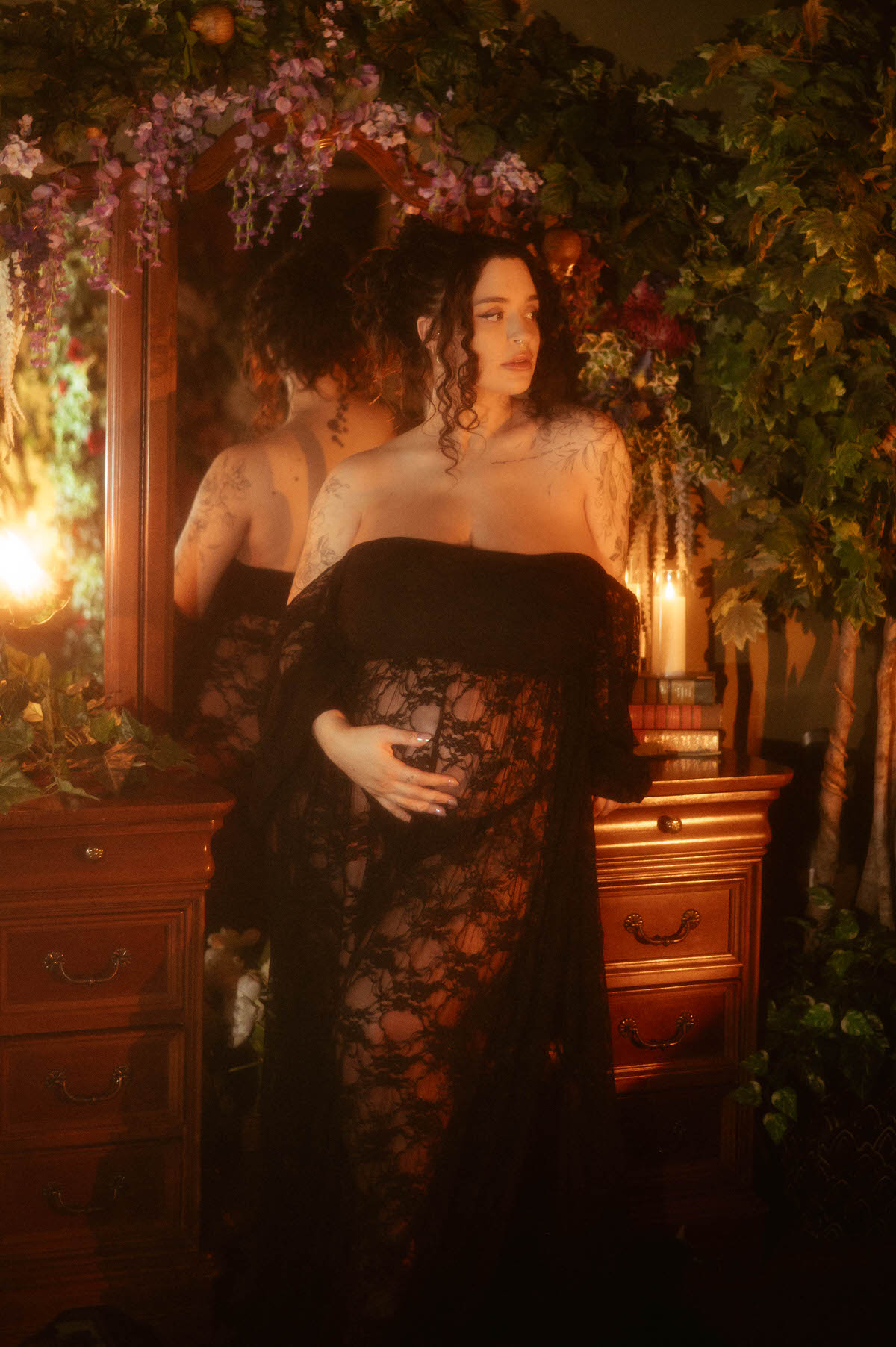 This maternity portrait studio Dallas image shows a woman standing in front of an antique wood dresser and mirror, one hand on her belly, wearing a black lace gown beneath hanging wisteria blooms at Royal Lune Photo.