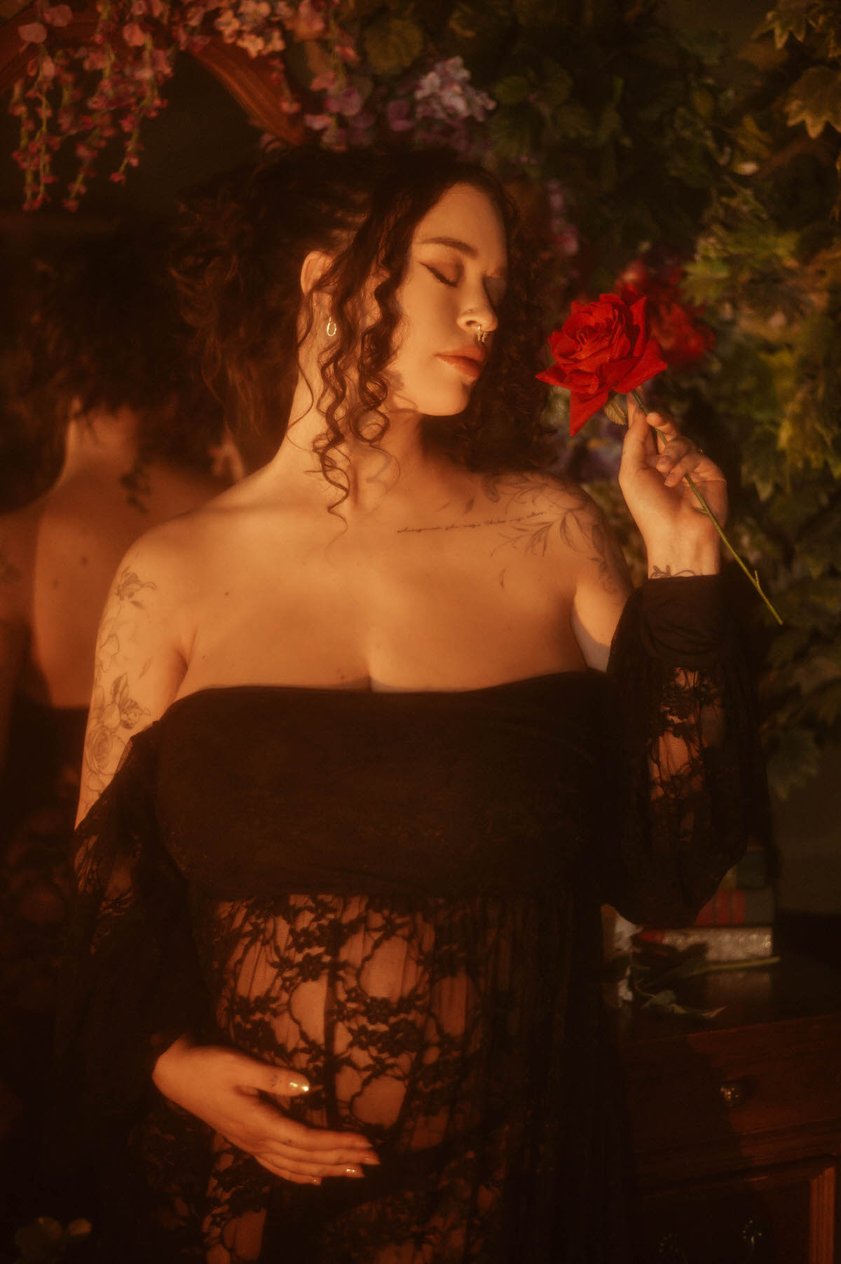 A Dallas maternity photographer captures a woman with her eyes closed holding a single red rose near her face, wearing an off the shoulder black lace gown with long sheer sleeves against a floral backdrop at Royal Lune Photo.