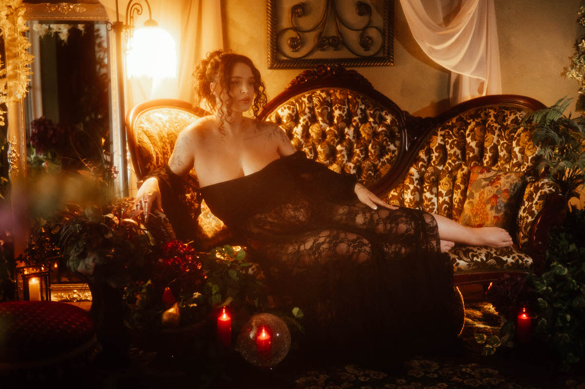 An artistic maternity photoshoot Dallas image shows a woman reclining on a vintage floral upholstered sofa in a black lace gown, one arm stretched along the back cushions, surrounded by candlelight and greenery at Royal Lune Photo.