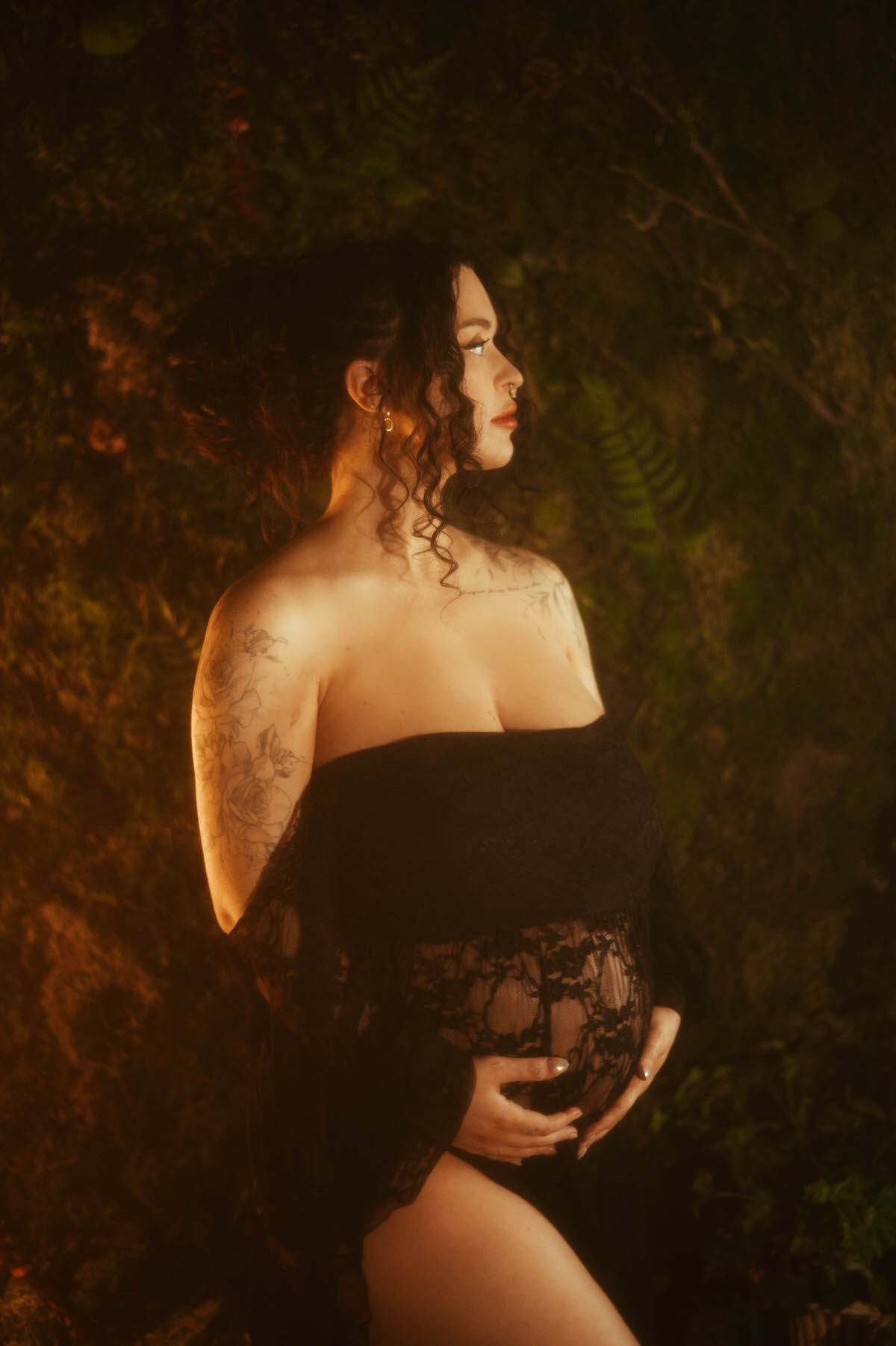 A moody maternity photography portrait captures a woman in profile wearing an off the shoulder black sheer lace gown, one hand resting on her belly, floral shoulder and arm tattoo work visible against a dark greenery backdrop at Royal Lune Photo.