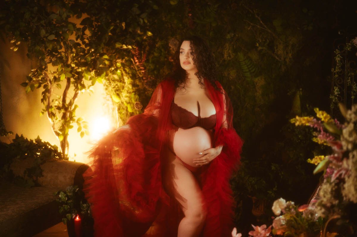 A Dallas maternity photographer captures a pregnant woman cradling her belly in a sheer red tulle robe, standing against a moss covered wall glowing with warm lantern light in the background at Royal Lune Photo.