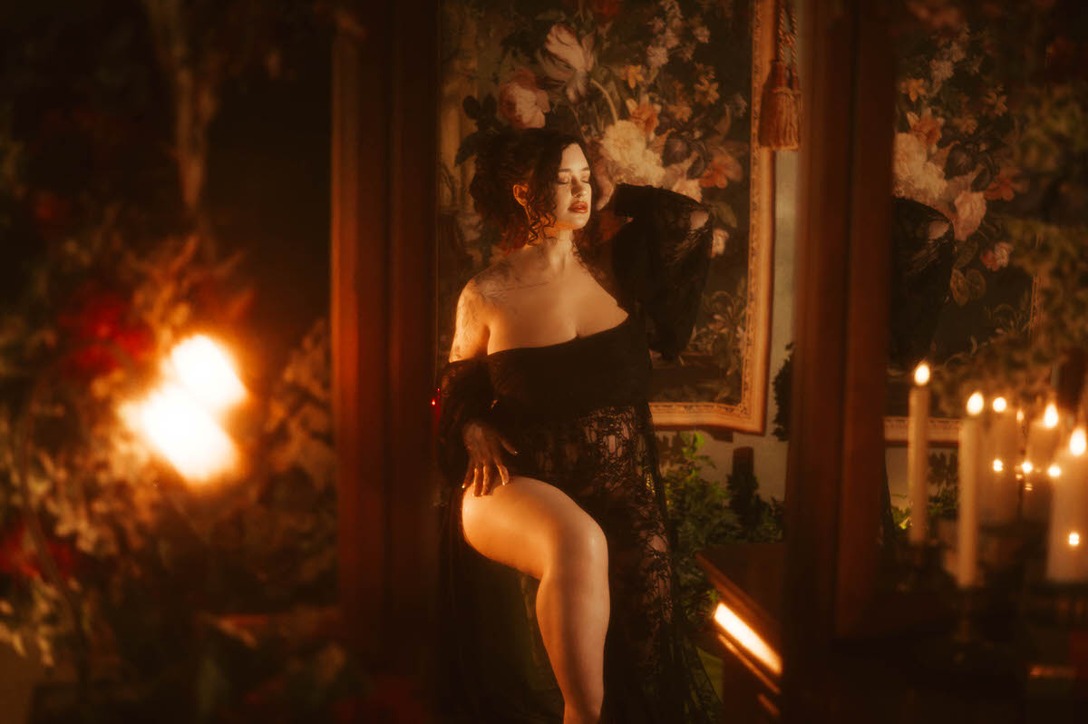 These pregnancy photoshoot ideas include a woman standing in profile with one leg propped on a ledge, wearing a black sheer lace gown open at the leg, lit by warm lamplight beside a floral tapestry at Royal Lune Photo.