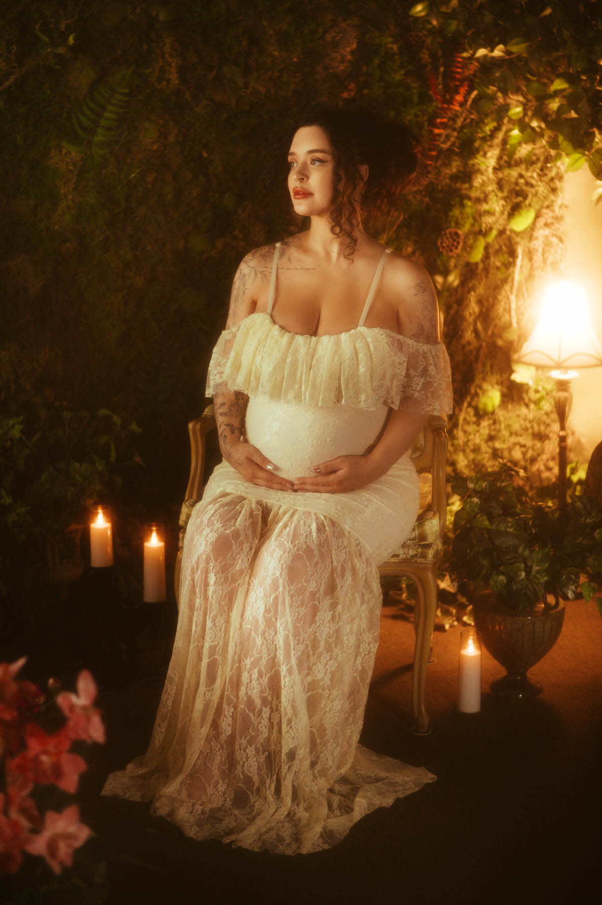 This maternity portrait studio Dallas image shows a woman seated in a gold vintage armchair wearing an ivory lace gown with a ruffled neckline, both hands clasped over her belly, framed by candlelight and blush florals at Royal Lune Photo.