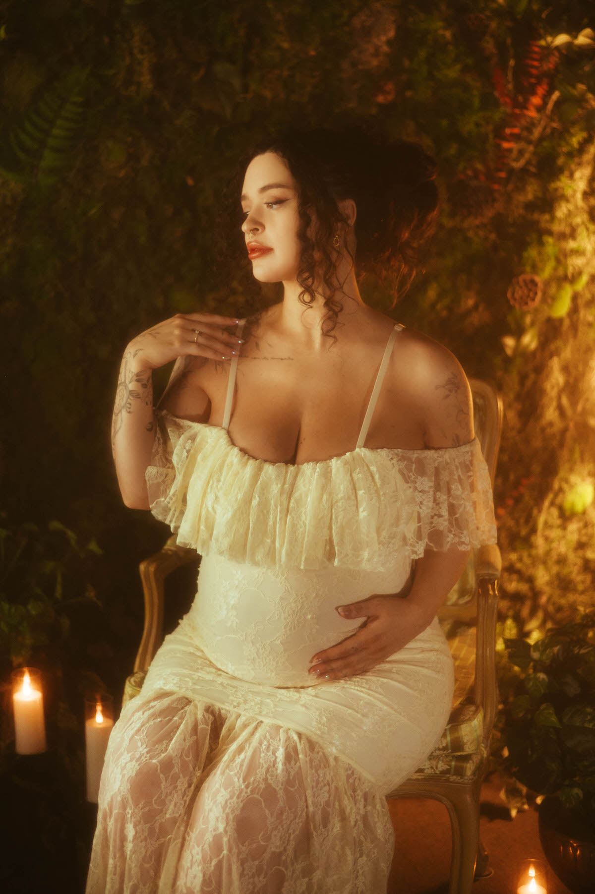 An artistic maternity photoshoot Dallas session features a woman seated in a gold armchair with one hand raised near her collarbone and the other resting on her belly, wearing an ivory lace gown against a mossy green backdrop at Royal Lune Photo.