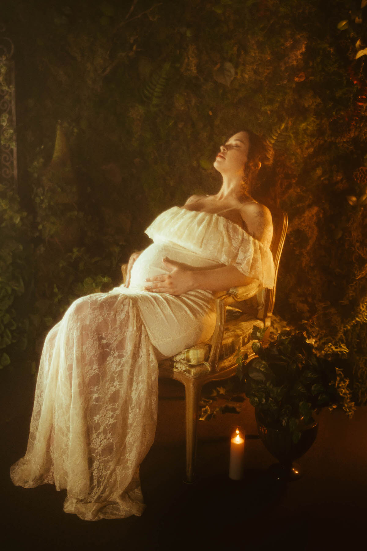 These pregnancy photoshoot ideas include a woman reclining in a gold vintage armchair with her head tilted back and eyes closed, wearing an ivory lace gown with one hand on her belly beside a lit candle at Royal Lune Photo.