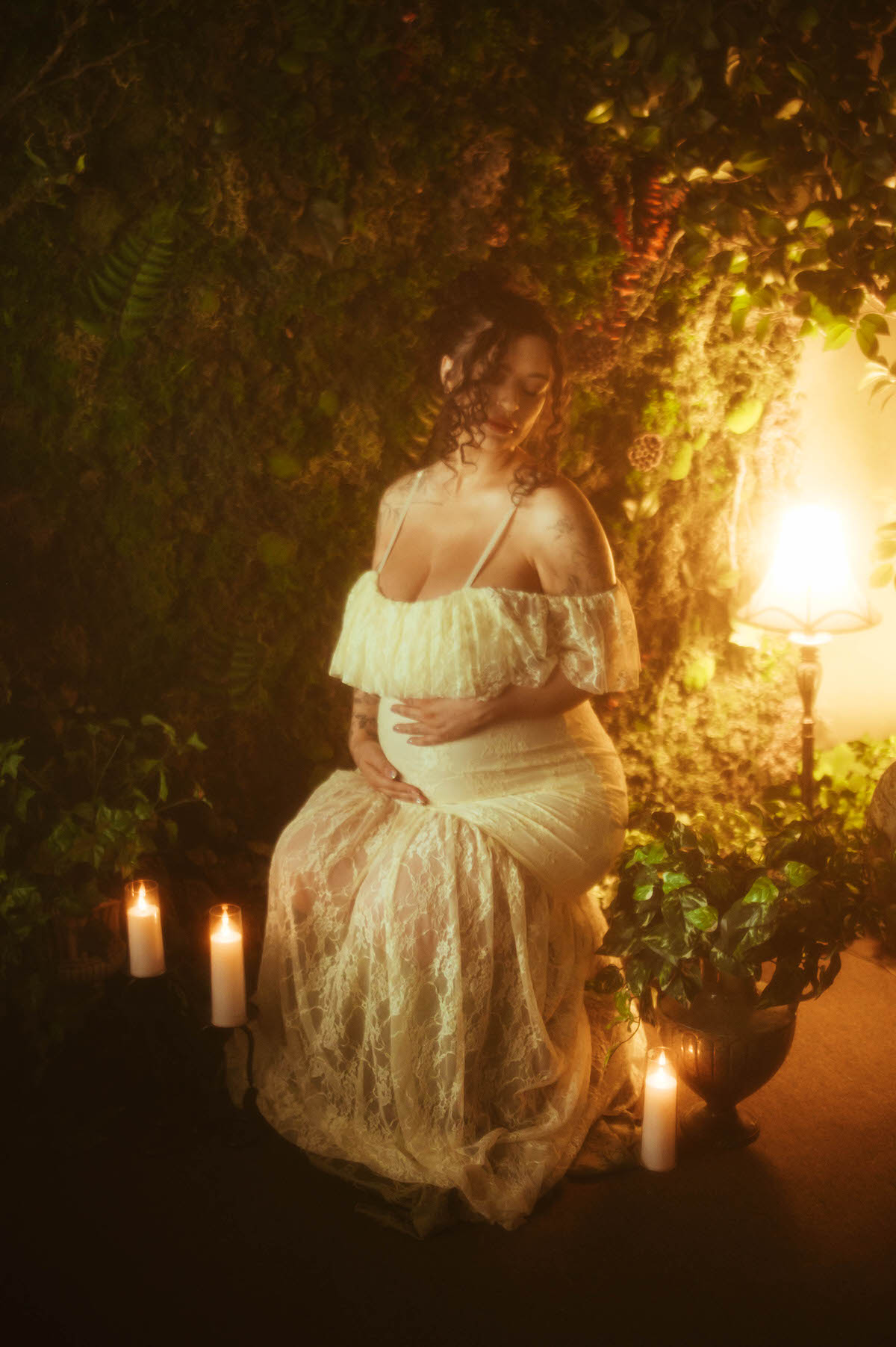 A Dallas maternity photographer captures a woman seated among greenery in an ivory lace gown with thin spaglietti straps, hand resting on her belly, flanked by pillar candles and warm lamplight at Royal Lune Photo.