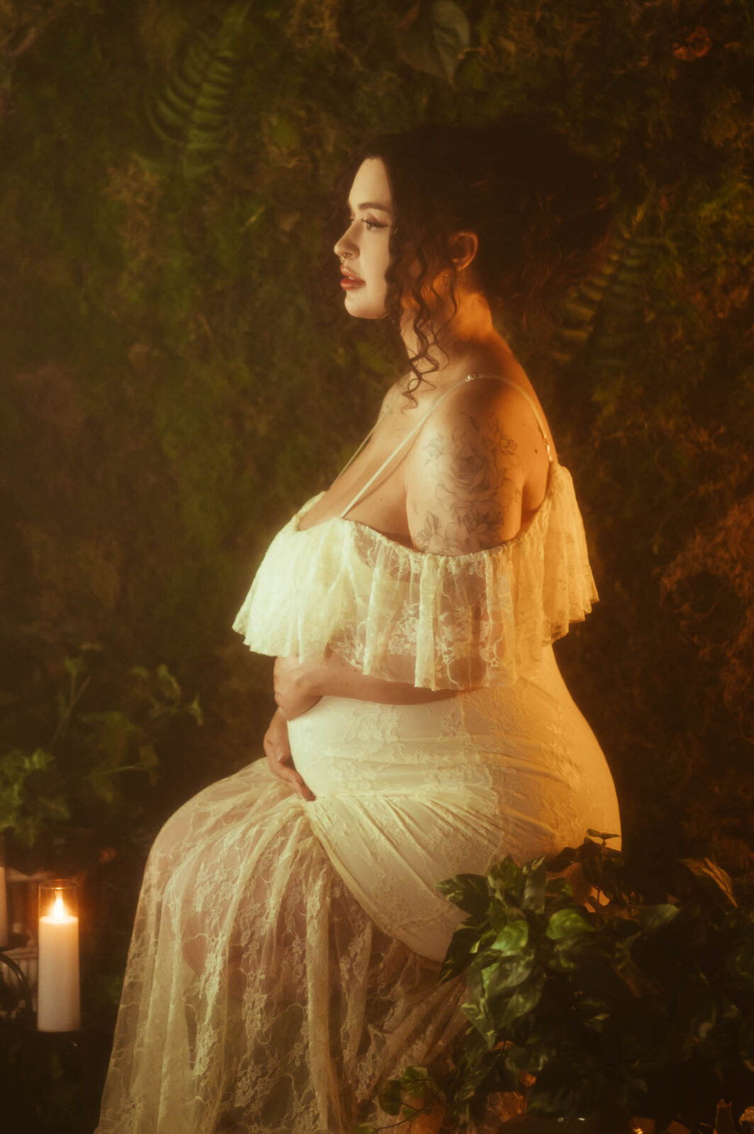 An artistic maternity photoshoot Dallas image shows a woman in profile wearing an ivory lace off the shoulder gown with a ruffled neckline, one hand resting on her belly, seated against a mossy backdrop lit by a single candle at Royal Lune Photo.