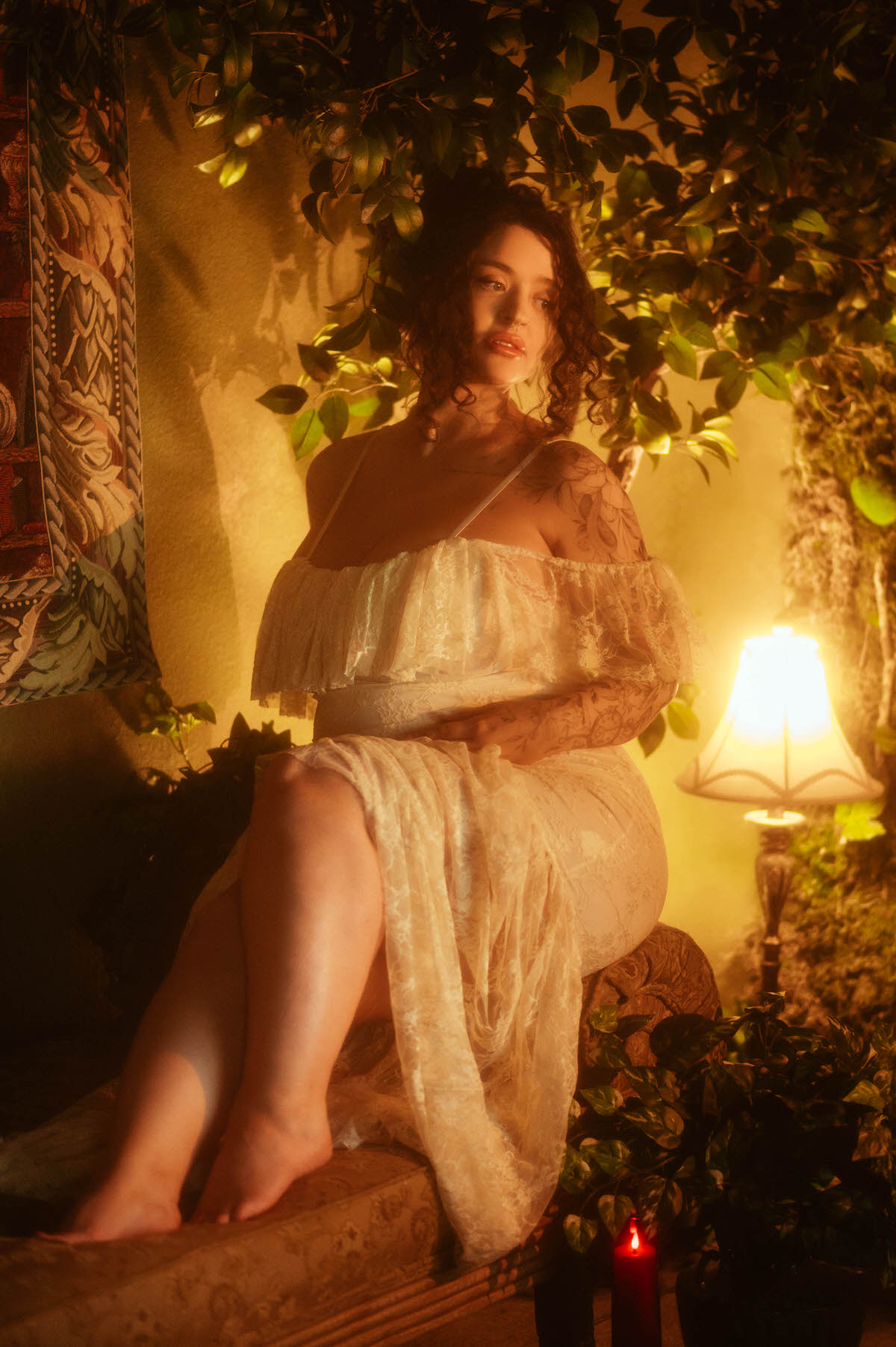 An artistic maternity photoshoot Dallas image shows a woman seated on a tufted bench in an ivory lace gown, one hand resting on her belly, leaning against a tapestry wall hanging beside a lit lamp at Royal Lune Photo.