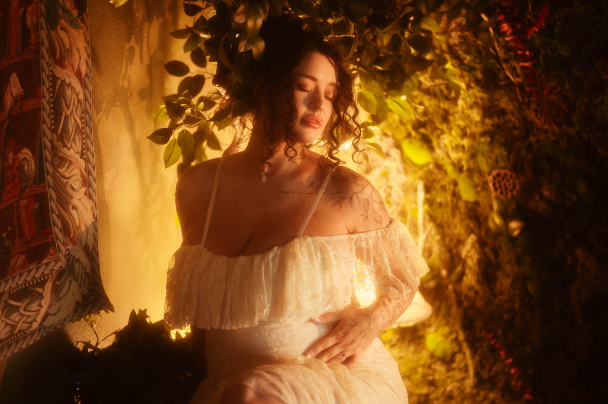 A close up moody maternity photography image shows a woman with her eyes closed wearing an ivory lace off the shoulder gown, one hand resting on her belly against a backdrop of greenery and warm lamplight at Royal Lune Photo.