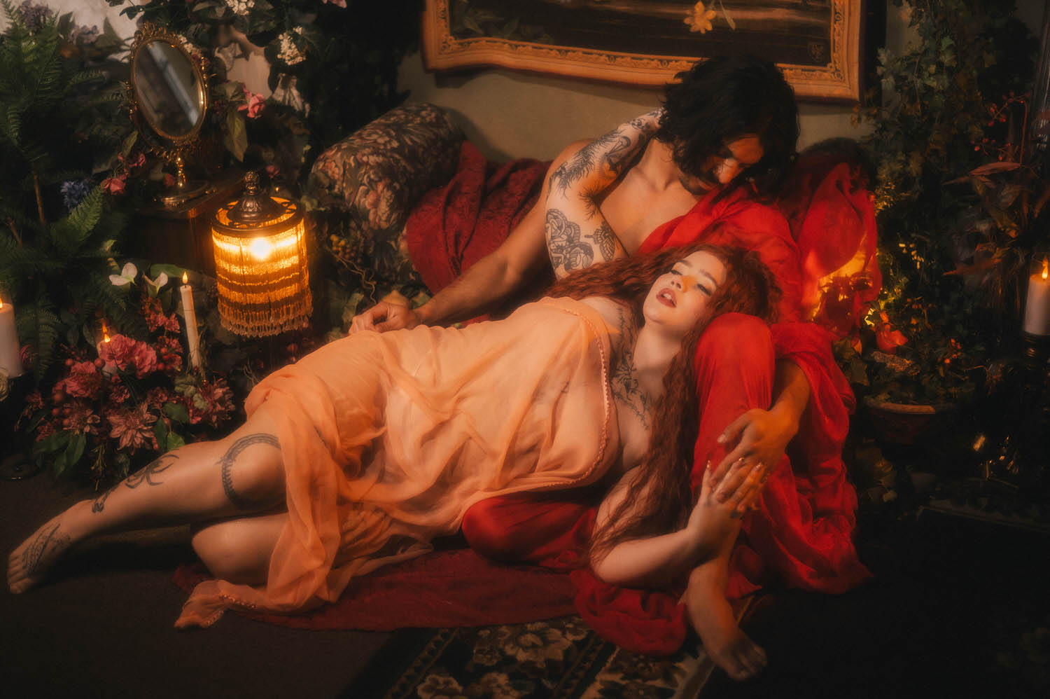 Renaissance boudoir photoshoot moment captured beside a warm fringed lamp, the woman in peach silk resting against her partner draped in wet red fabric. Royal Lune Photo