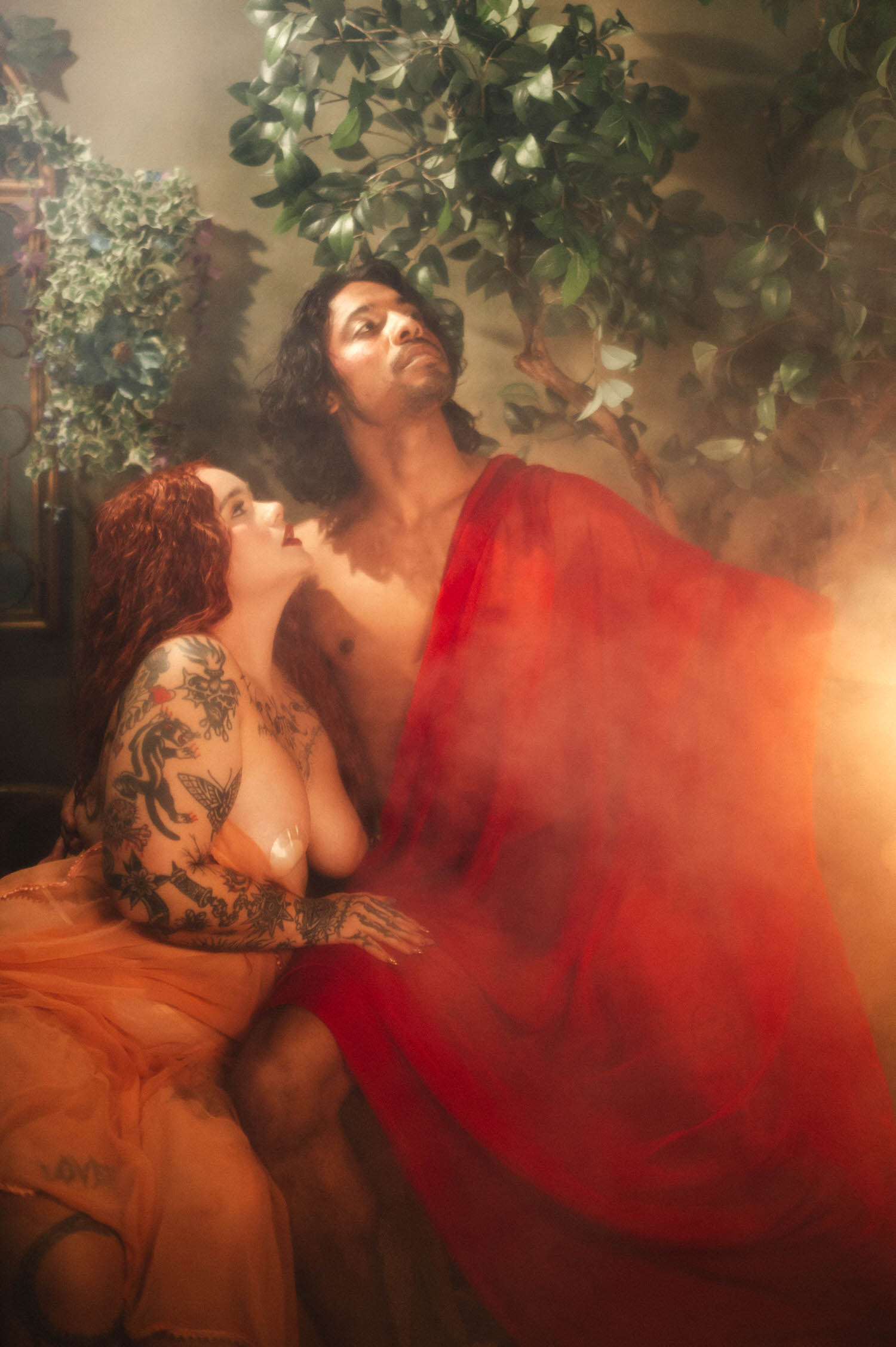 Dallas Fort Worth boudoir photographer captures the couple gazing skyward together, the man's wet red drape catching golden light beneath green foliage. Royal Lune Photo