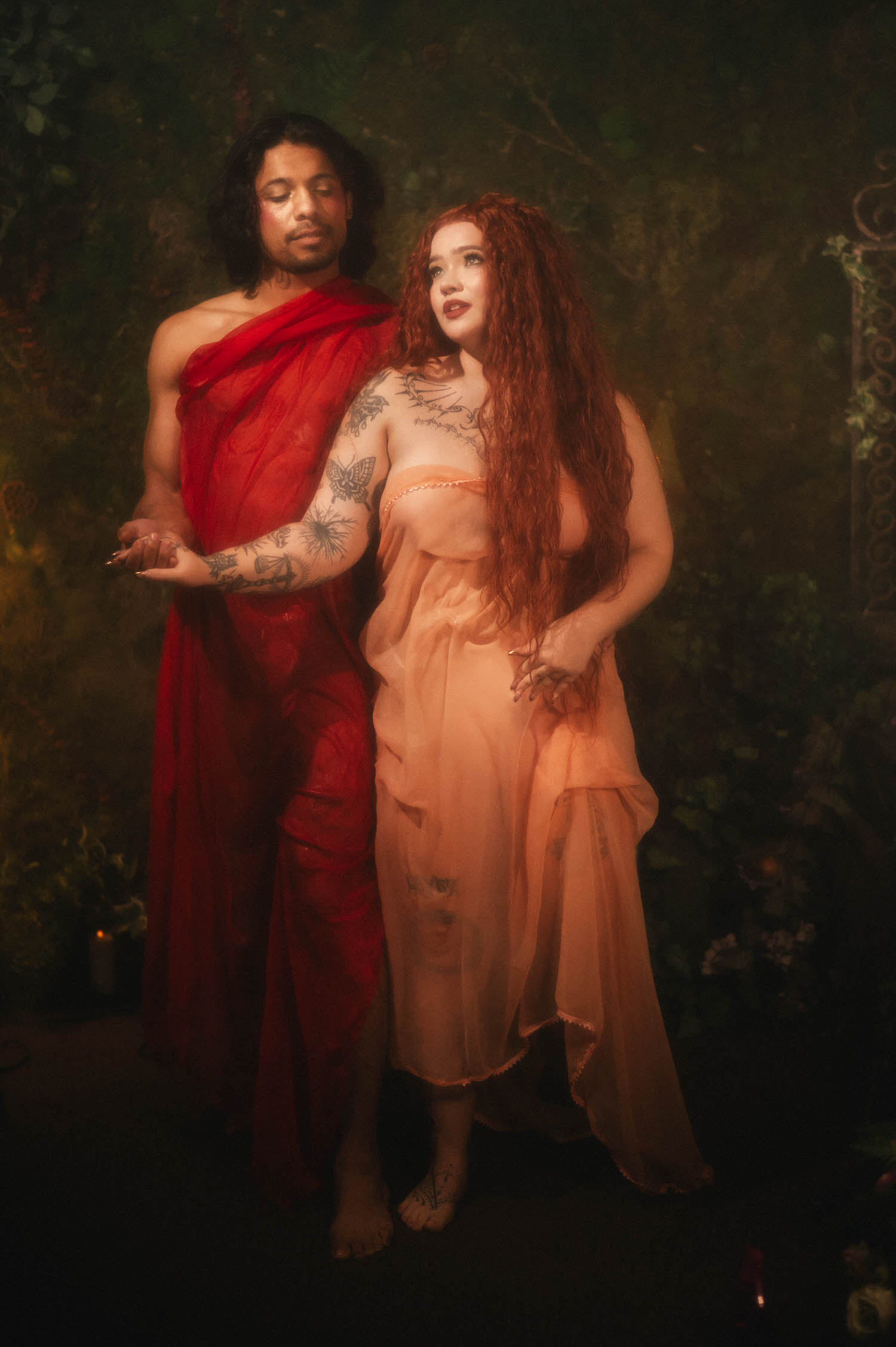 Romantic couples photography Texas portrait captures the couple standing close in flowing peach and red silk, deep green foliage filling the background. Royal Lune Photo