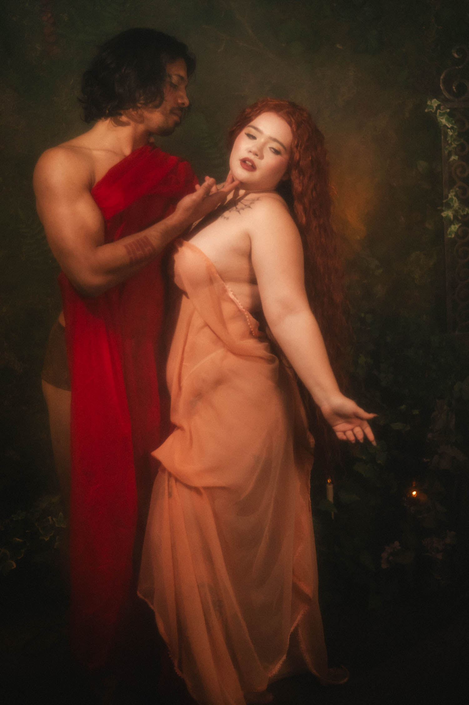 A painterly couples boudoir portrait inspired by renaissance paintings captures the woman glancing back over her bare shoulder, her partner's hand at her throat amid dark garden greenery. Royal Lune Photo